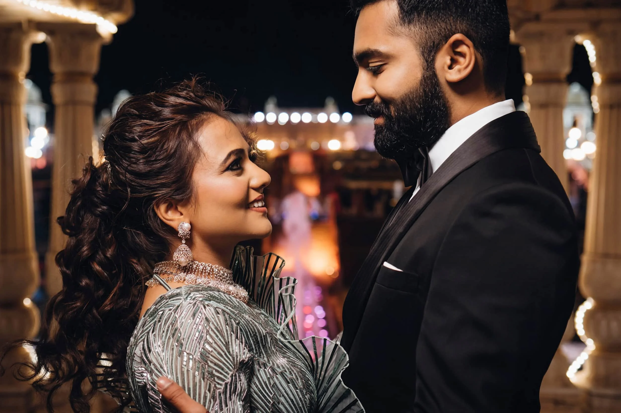 A couple dressed in formal attire, looking at each other lovingly in a romantic setting with warm lighting and decorative columns.
