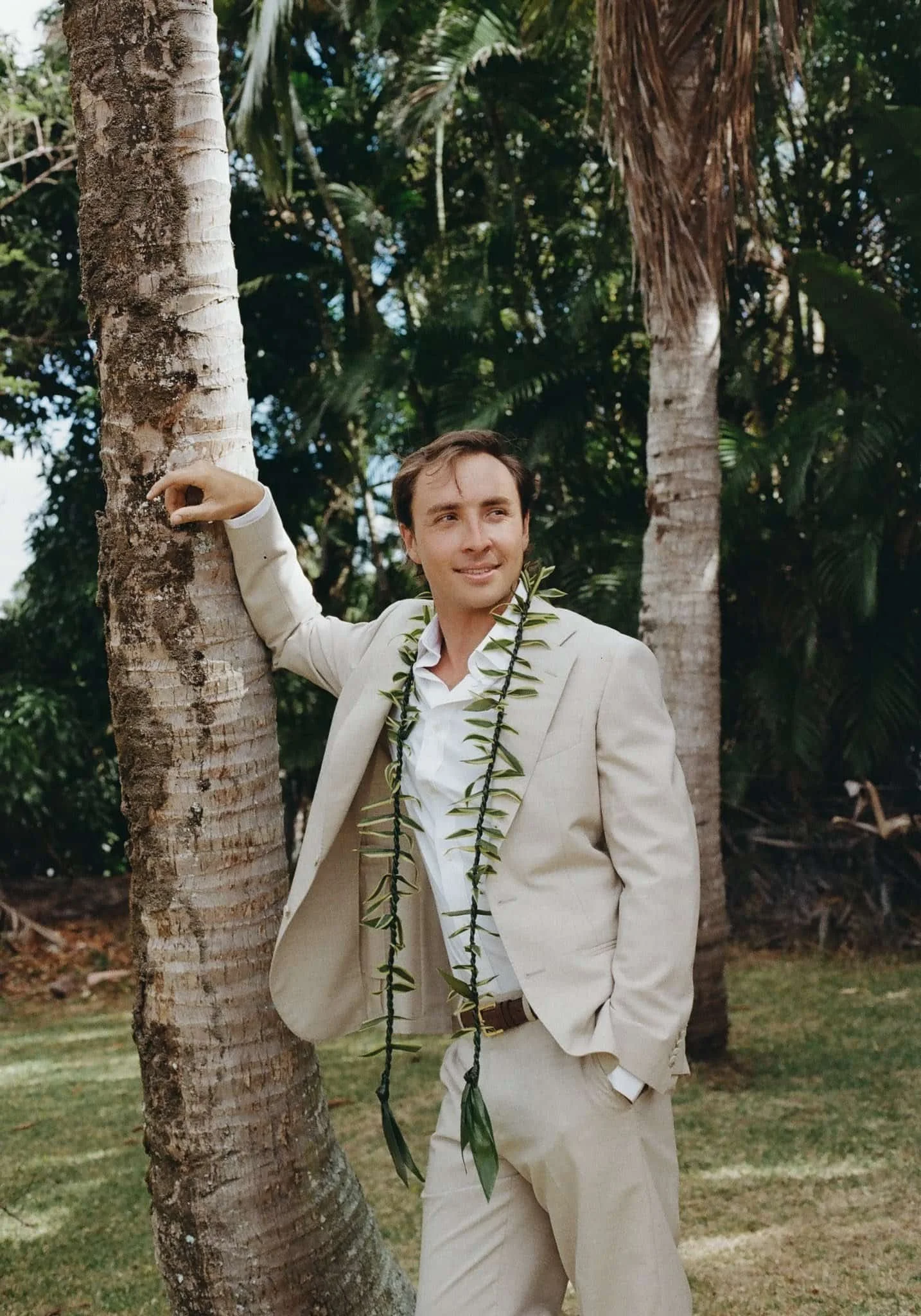 A man in a beige suit with a white shirt and a leafy lei around his neck is standing outdoors, leaning against a palm tree, smiling, in a lush green tropical setting.
