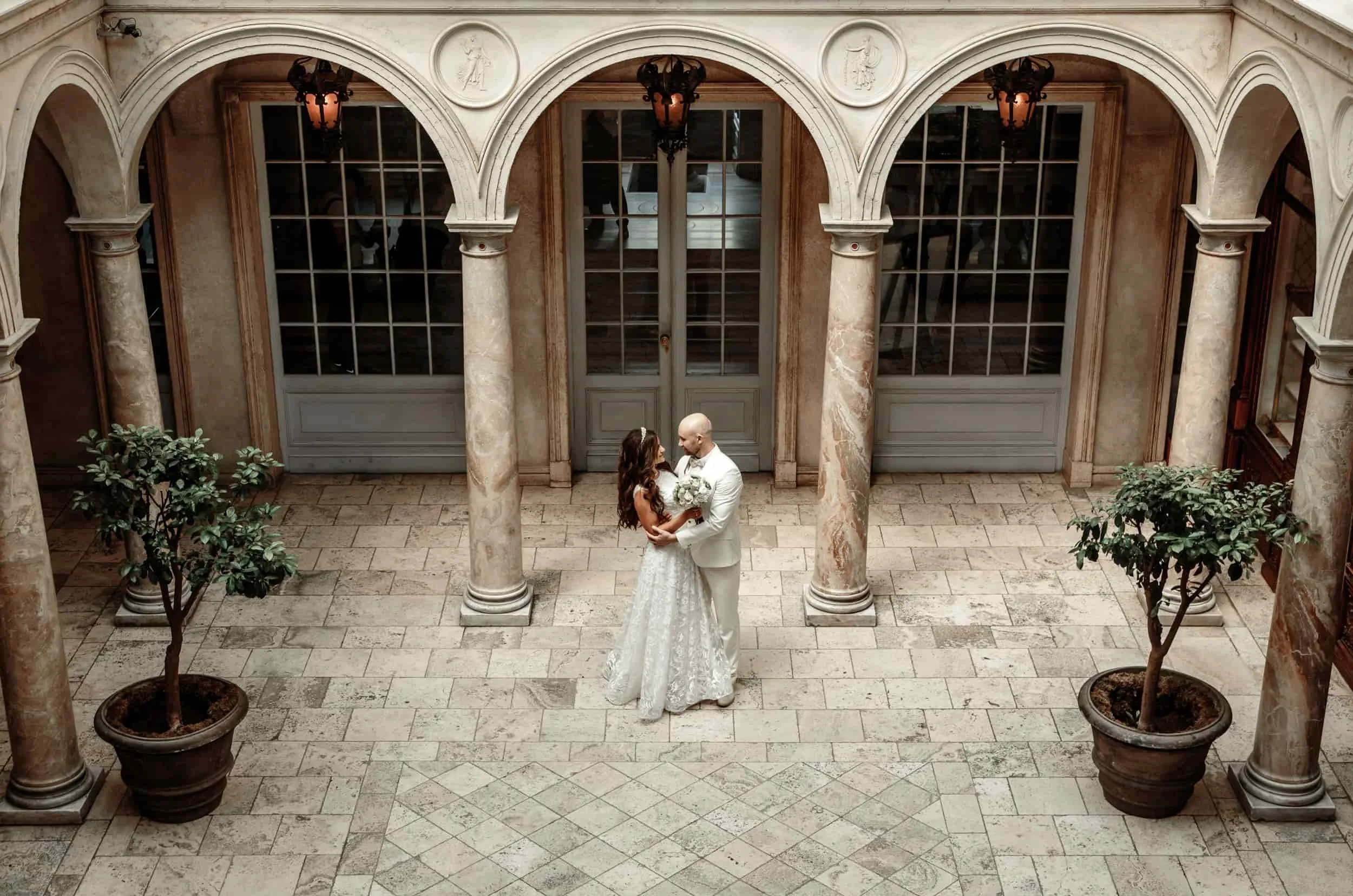 A bride and groom standing close together in an elegant, classical-style courtyard with marble columns, large windowed doors, and potted trees, sharing a moment.