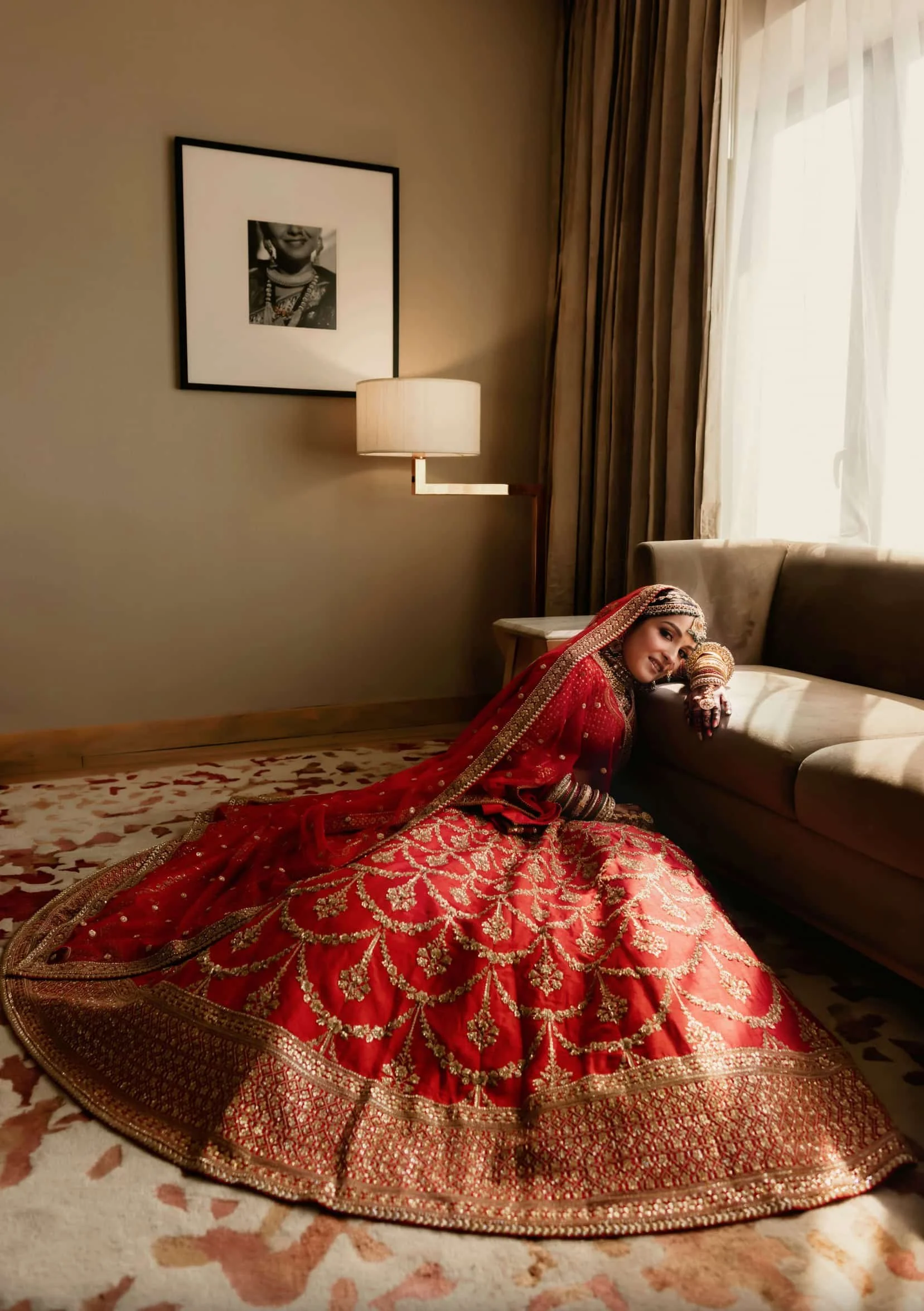 Woman in traditional red and gold Indian bridal dress lying on the floor with head resting on a sofa, in a room with a framed black and white photograph on the wall, a lamp, curtains, and a patterned rug.