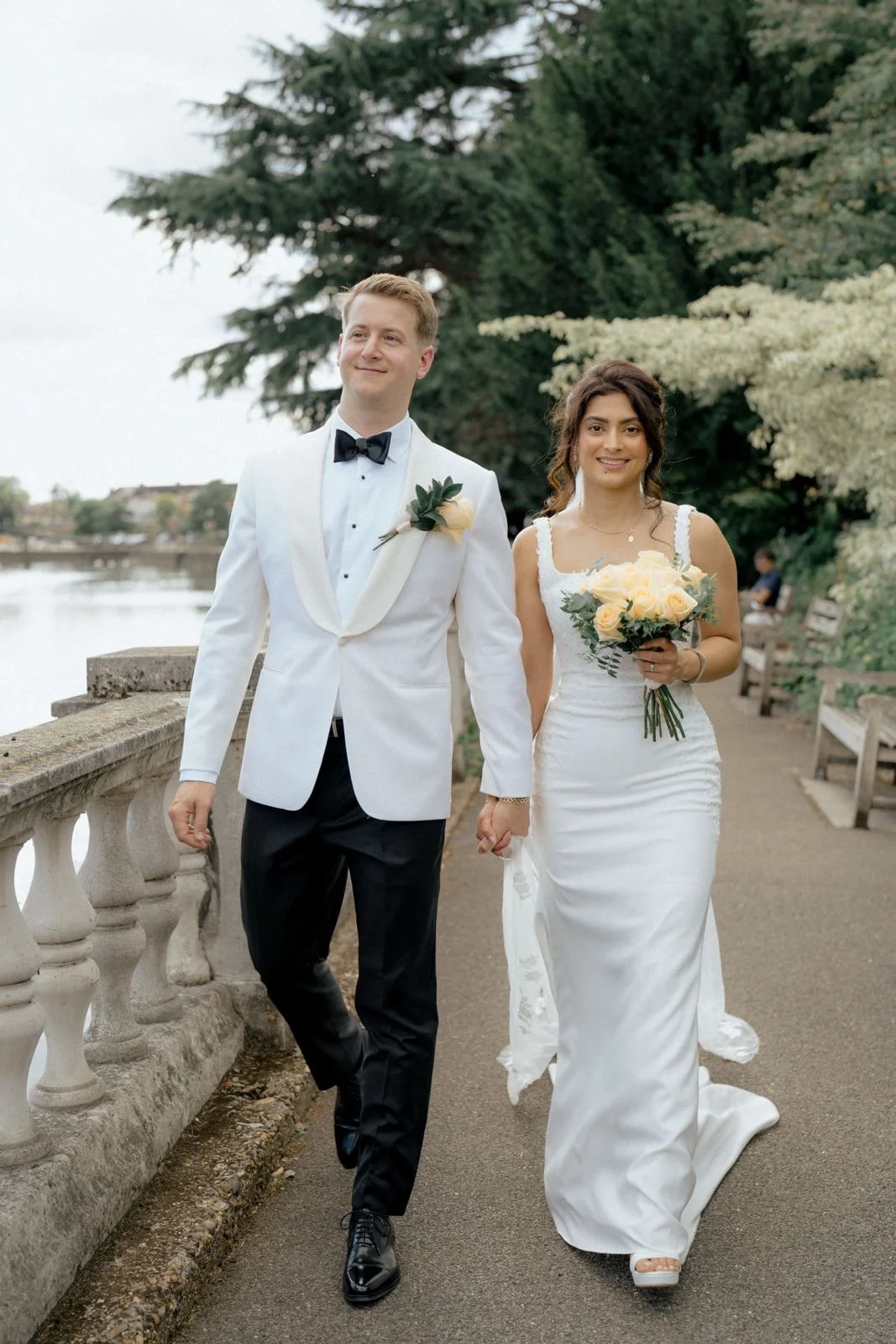 A bride and groom walking hand in hand outdoors near a body of water, with trees in the background. The groom is dressed in a white tuxedo jacket, black pants, and a black bow tie. The bride is wearing a white wedding gown and holding a bouquet of pa