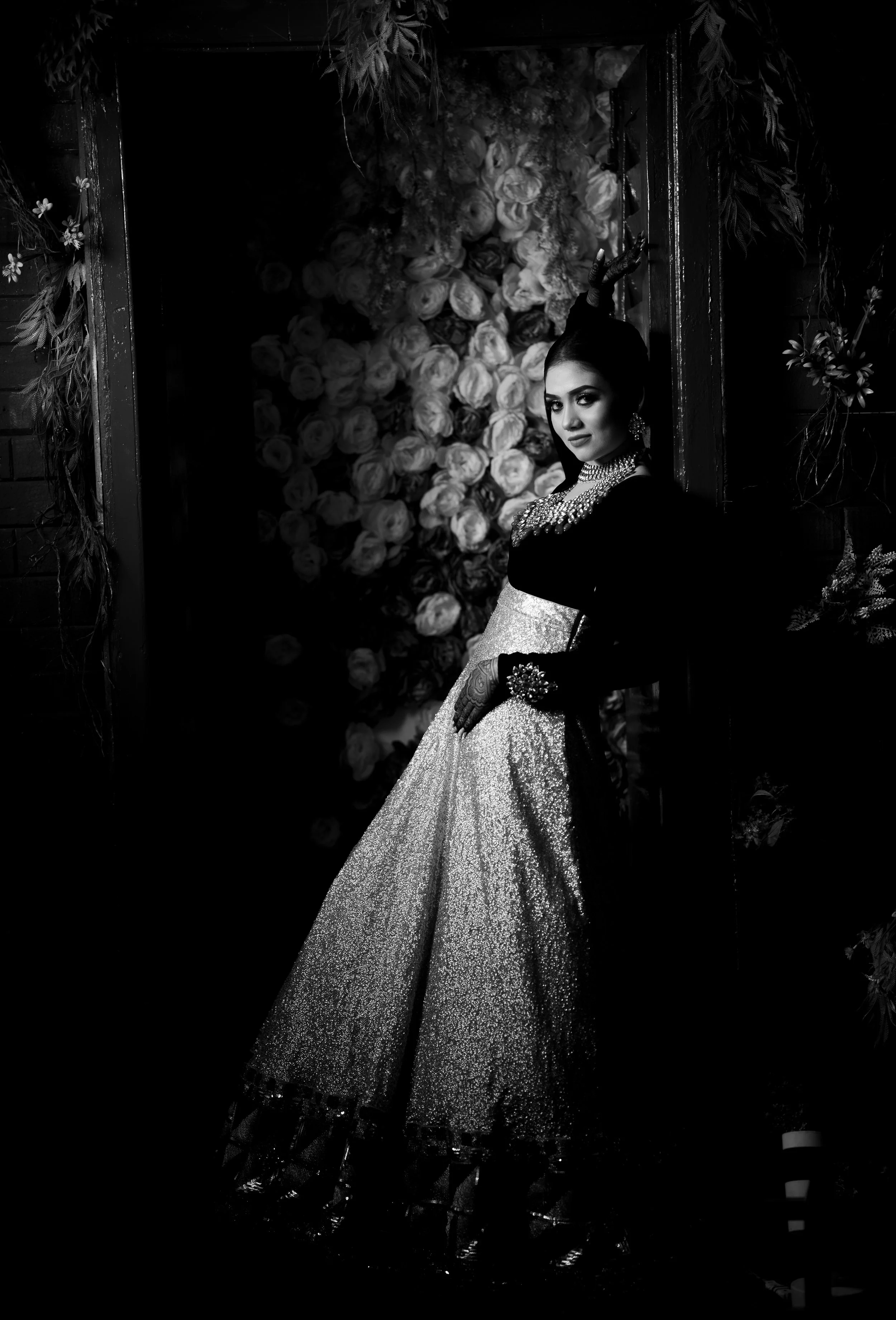 A woman dressed in traditional Indian attire, wearing jewelry, standing in front of a floral backdrop in a dimly lit setting.