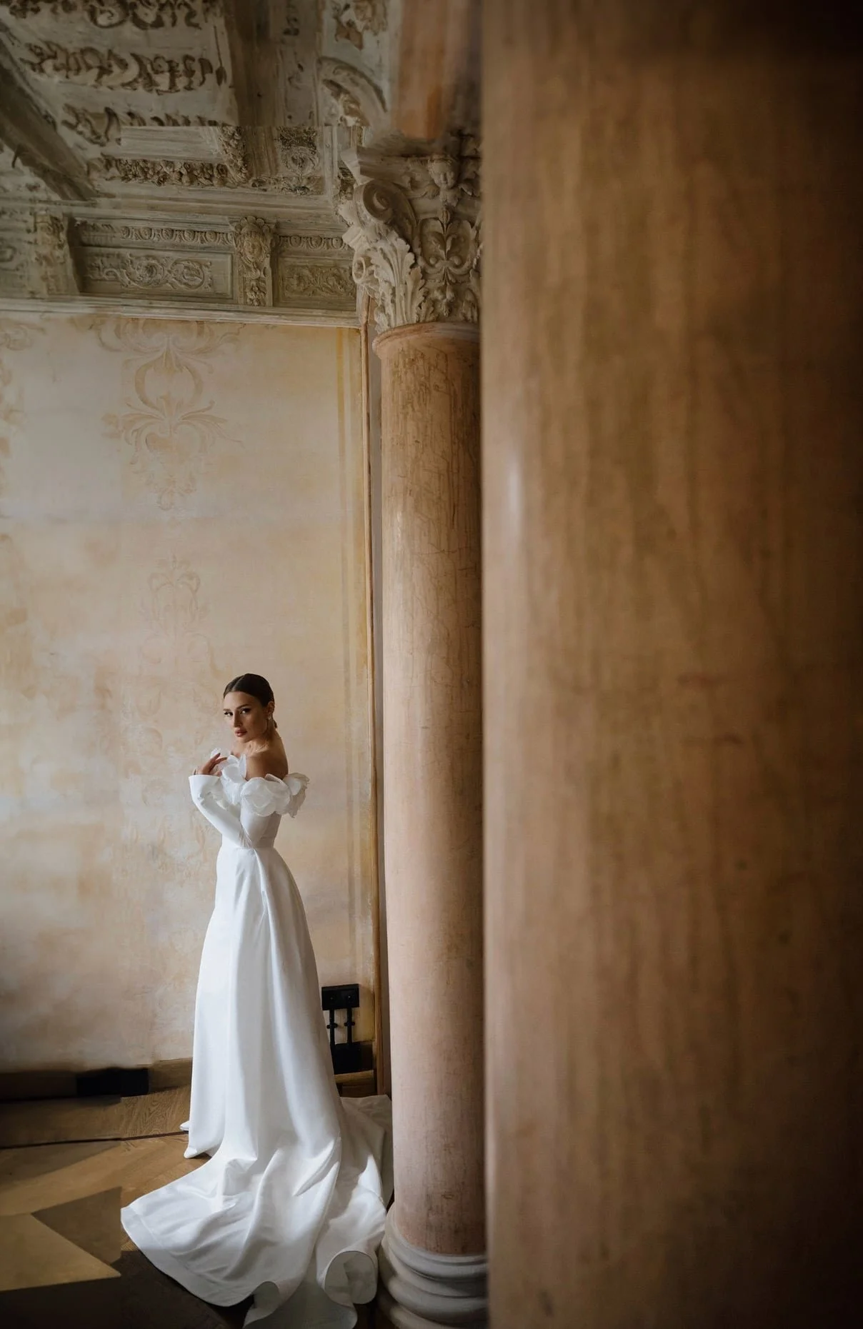 A woman in a long white dress with puffed sleeves standing in a historic, ornate room with decorative ceiling moldings and large stone columns.