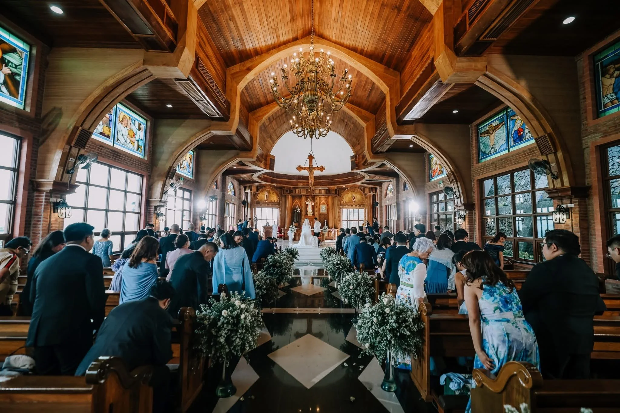 A church interior with a high wooden ceiling, stained glass windows, a chandelier, and an altar where a wedding ceremony is taking place. Guests are seated and standing, facing the altar.