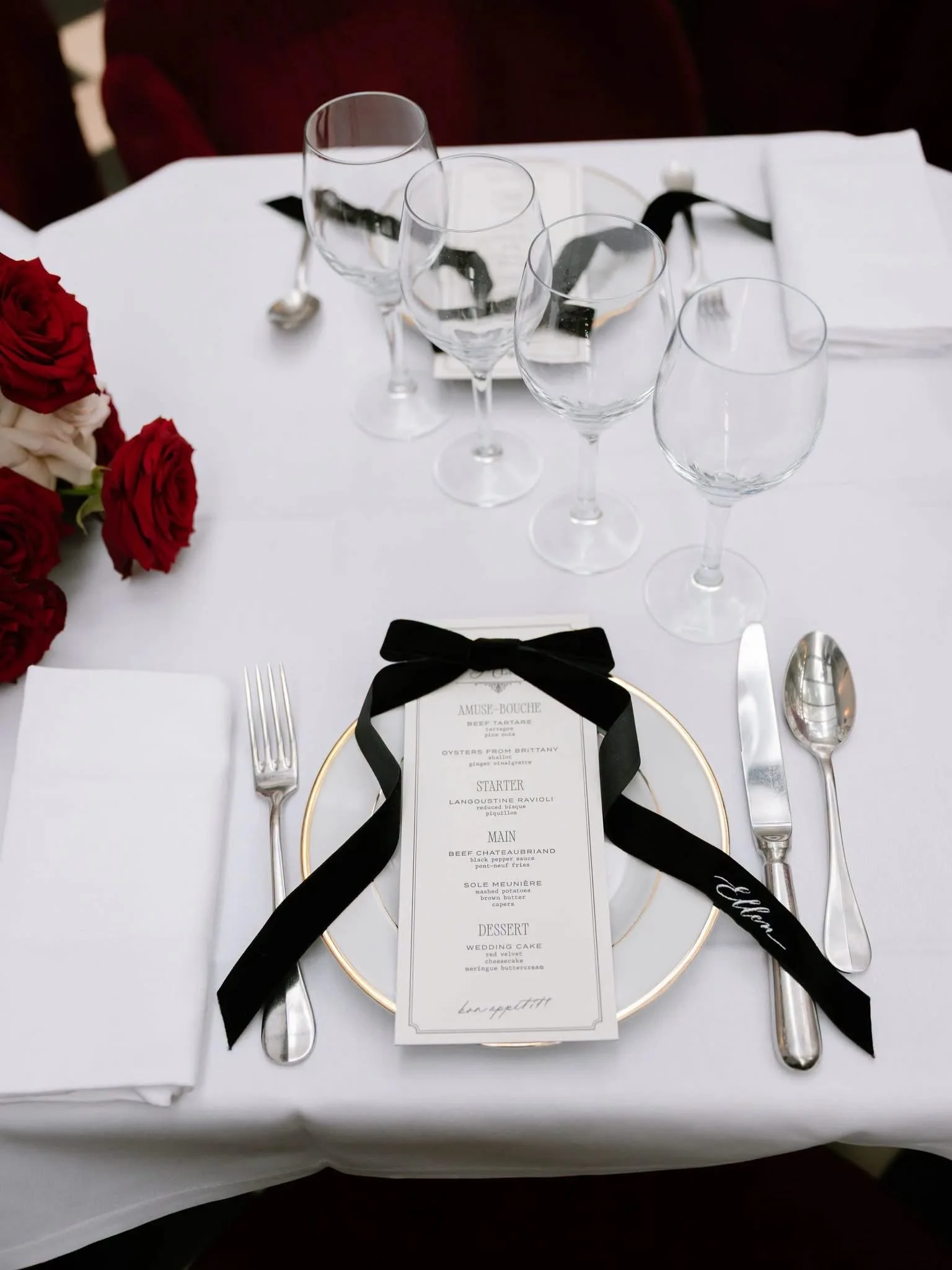 Elegant dining table setup for a wedding reception with a white tablecloth, empty wine glasses, silverware, a floral arrangement with red roses, and a menu card tied with a black ribbon.