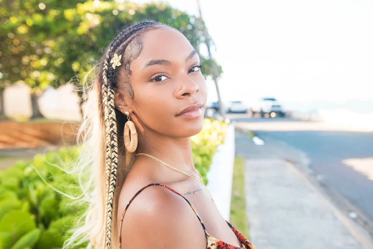 A young woman with long braided hair, hoop earrings, and a floral dress standing on a sidewalk near trees and parked cars, looking at the camera.
