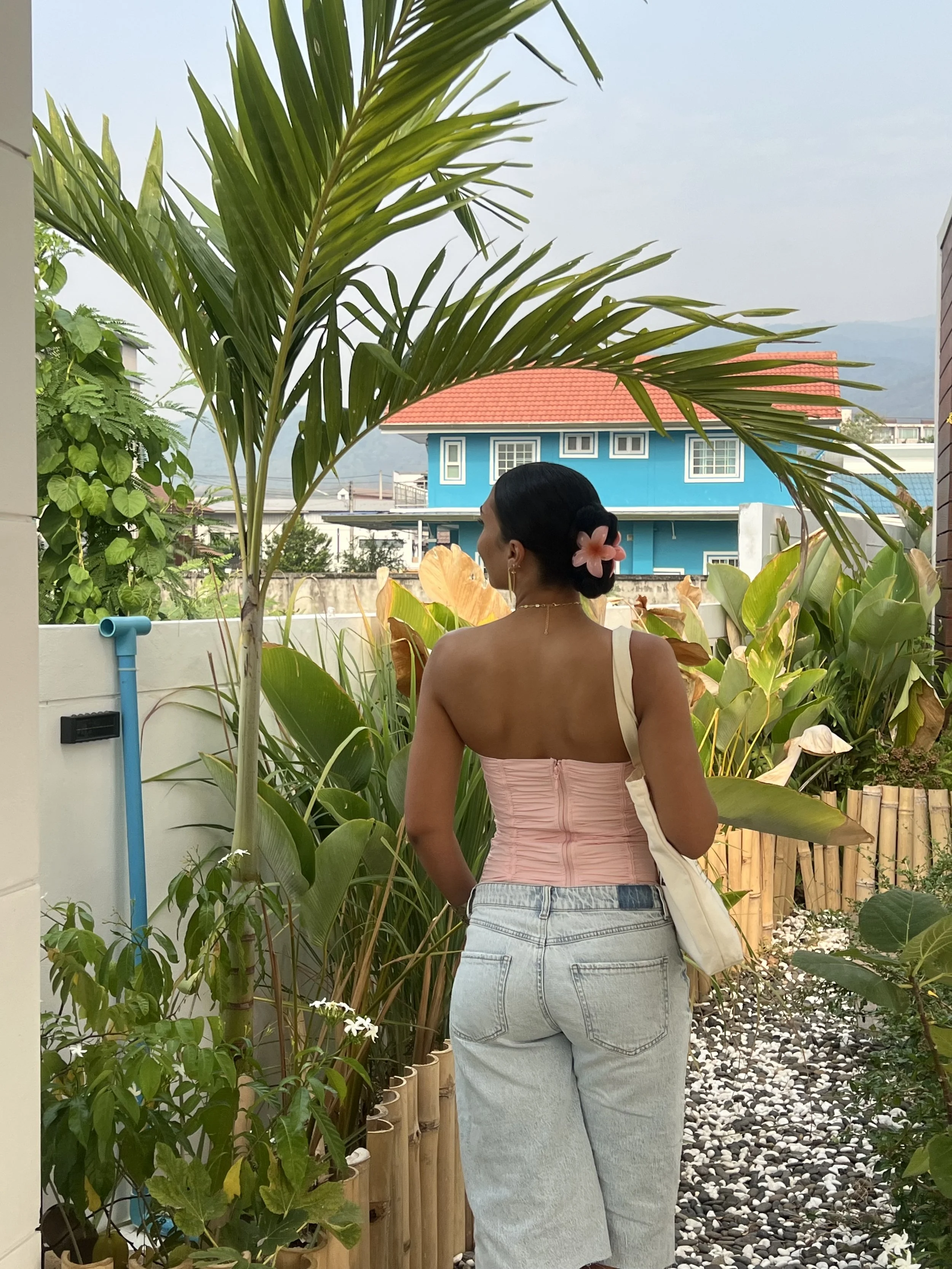 A woman with dark hair in a bun, decorated with a pink flower, stands among tropical plants in a garden, facing away, with a blue house with a red roof in the background.