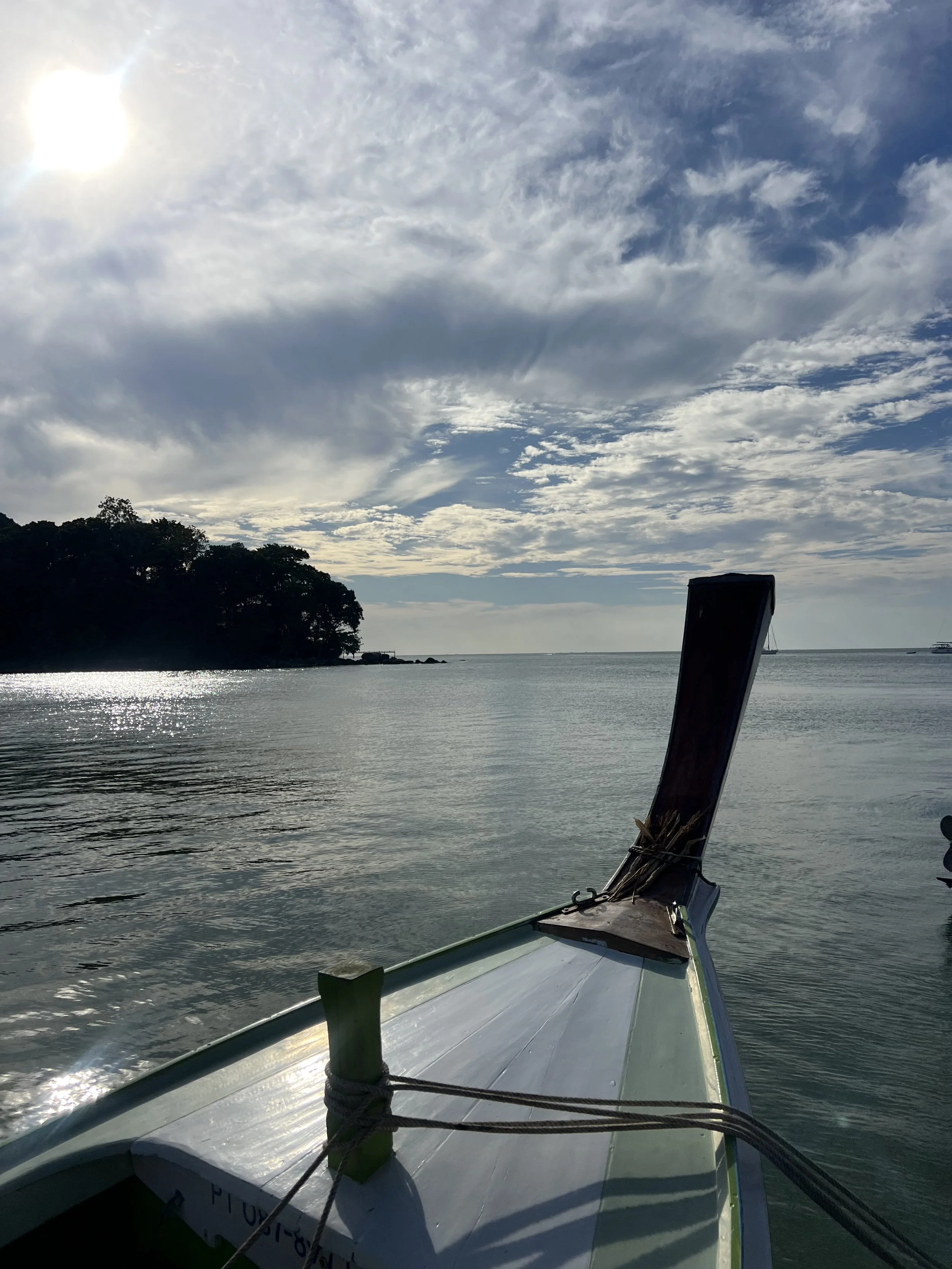 The image shows the front part of a boat on calm water, with a small island covered in trees on the left and some boats or ships visible in the distance. The sky is partly cloudy with the sun shining brightly.
