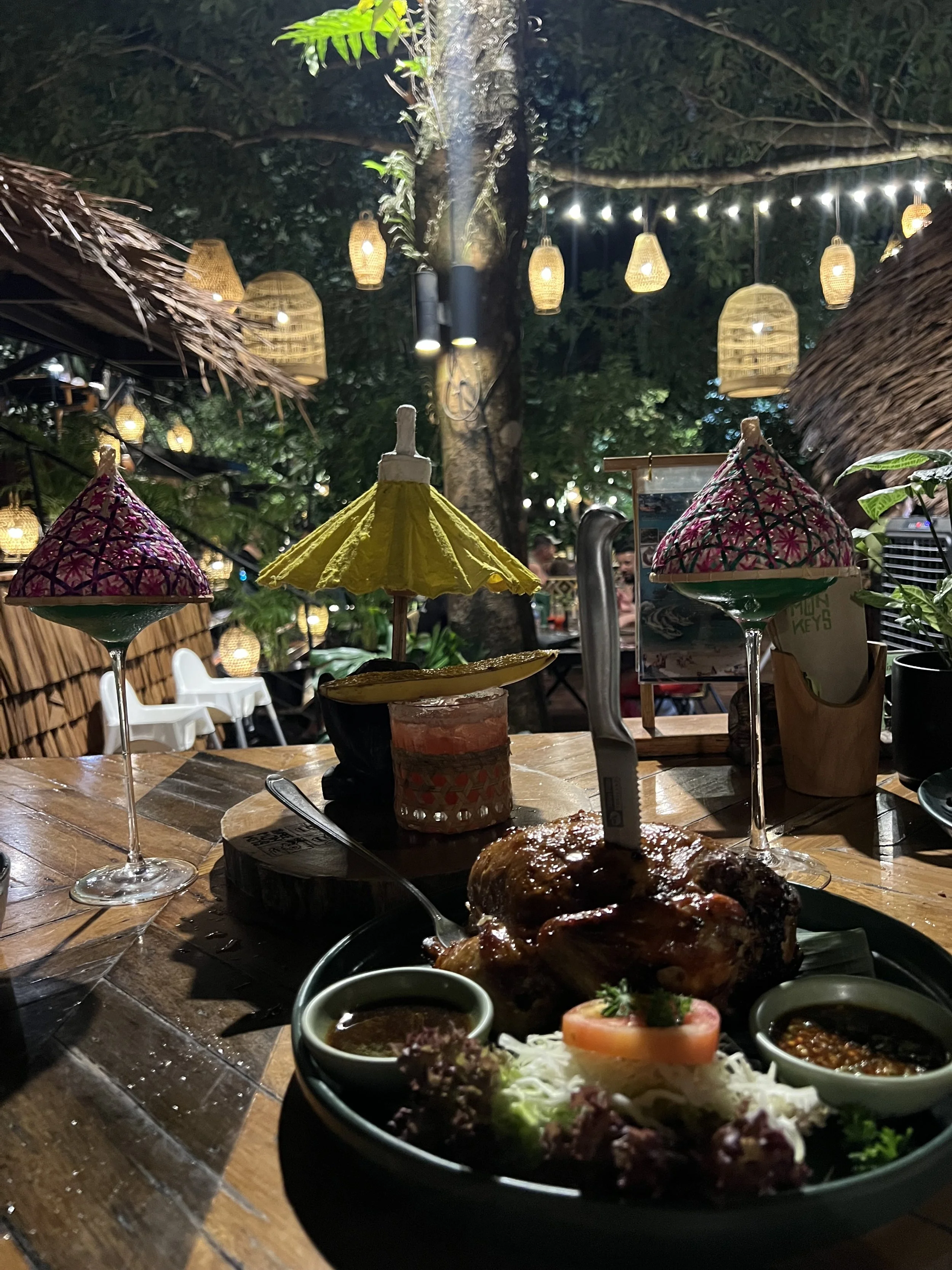 A meal on a wooden table at an outdoor restaurant during the evening, featuring a roasted chicken with dipping sauces, surrounded by small drink umbrellas, with decorative hanging lanterns and string lights illuminating the background.
