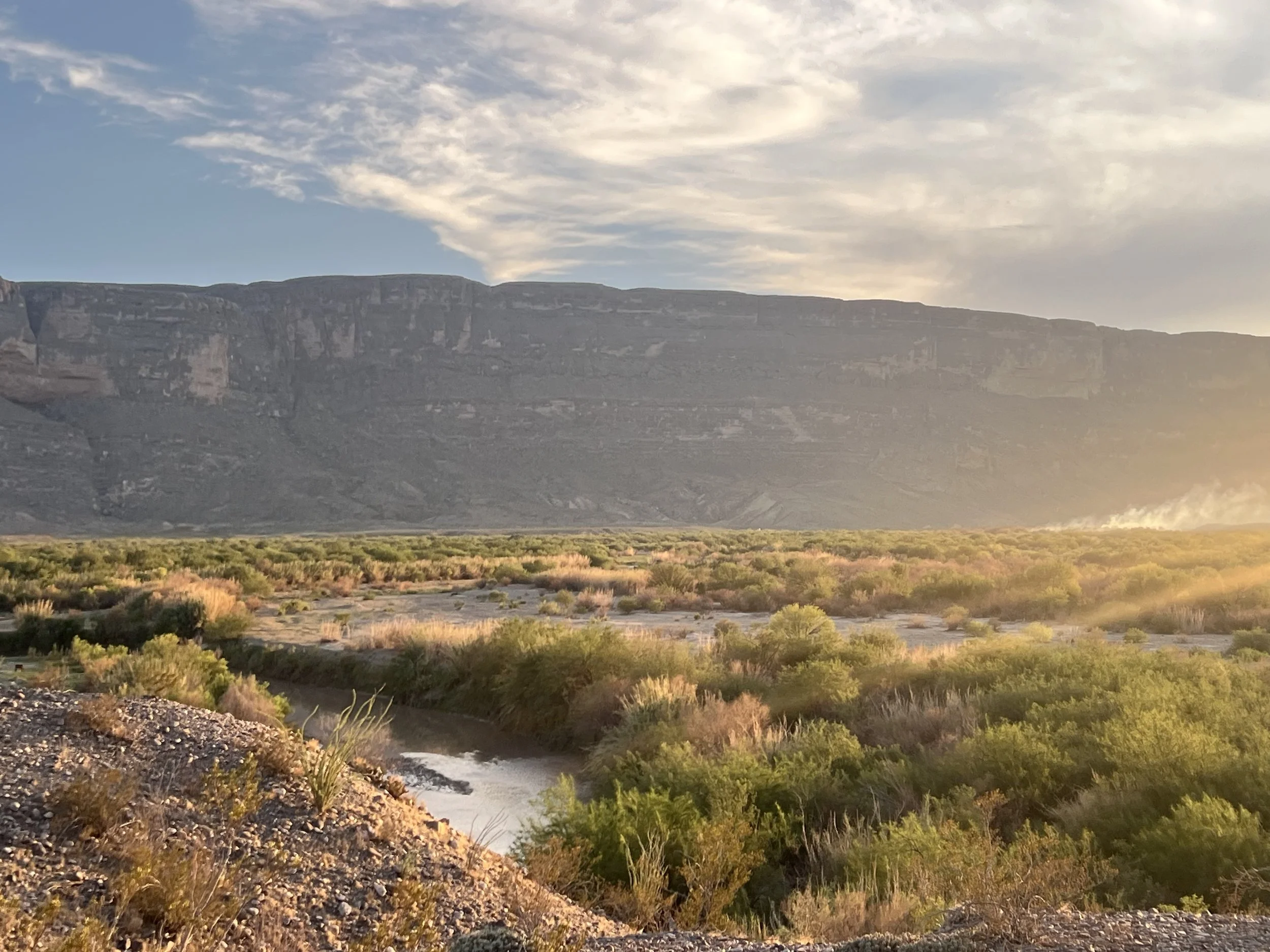 Sunset over a valley with a river, surrounded by green bushes and a mountain range in the background.