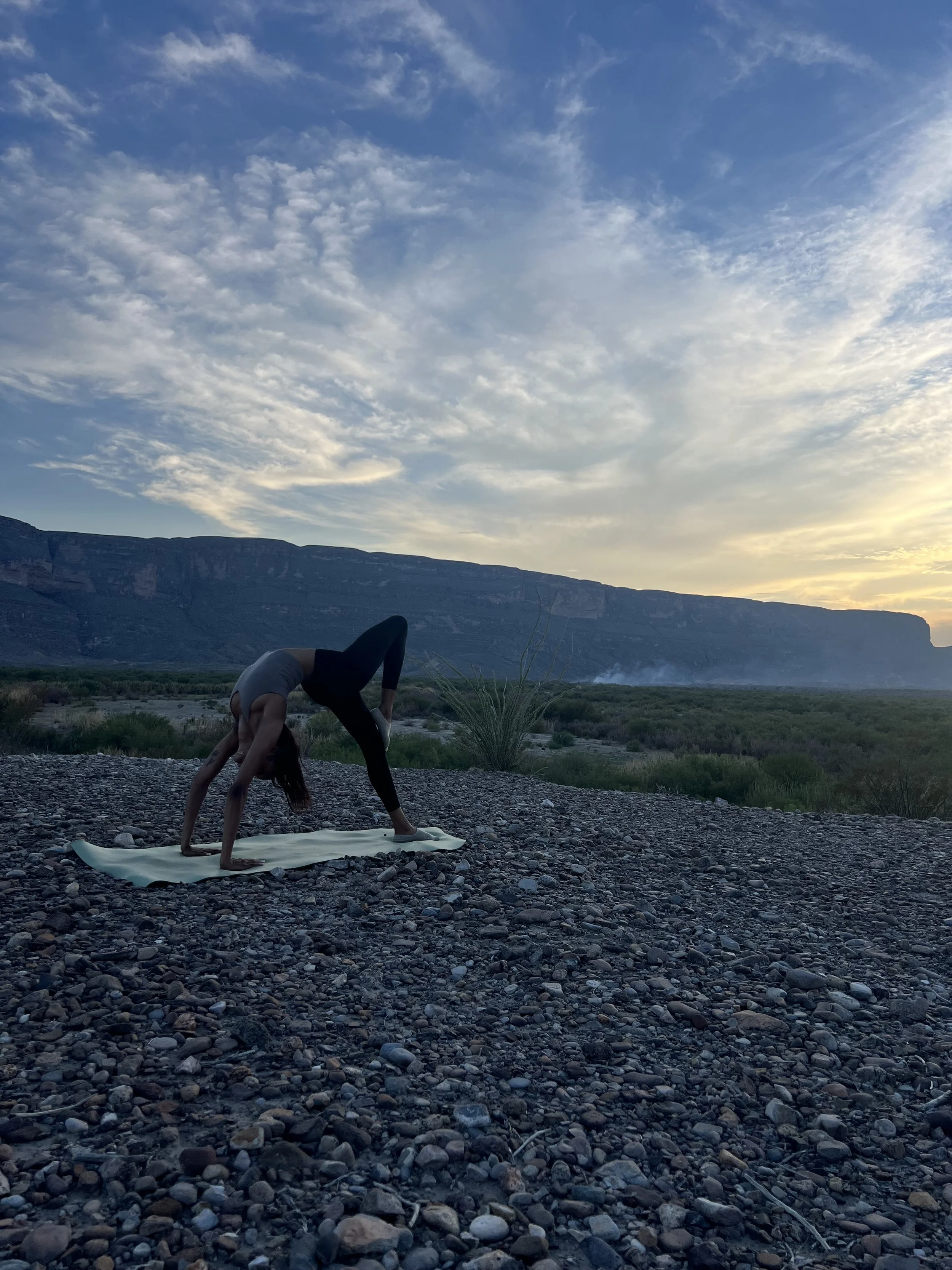 Person practicing yoga outdoors on a rocky terrain during sunset, with mountains and a partly cloudy sky in the background.