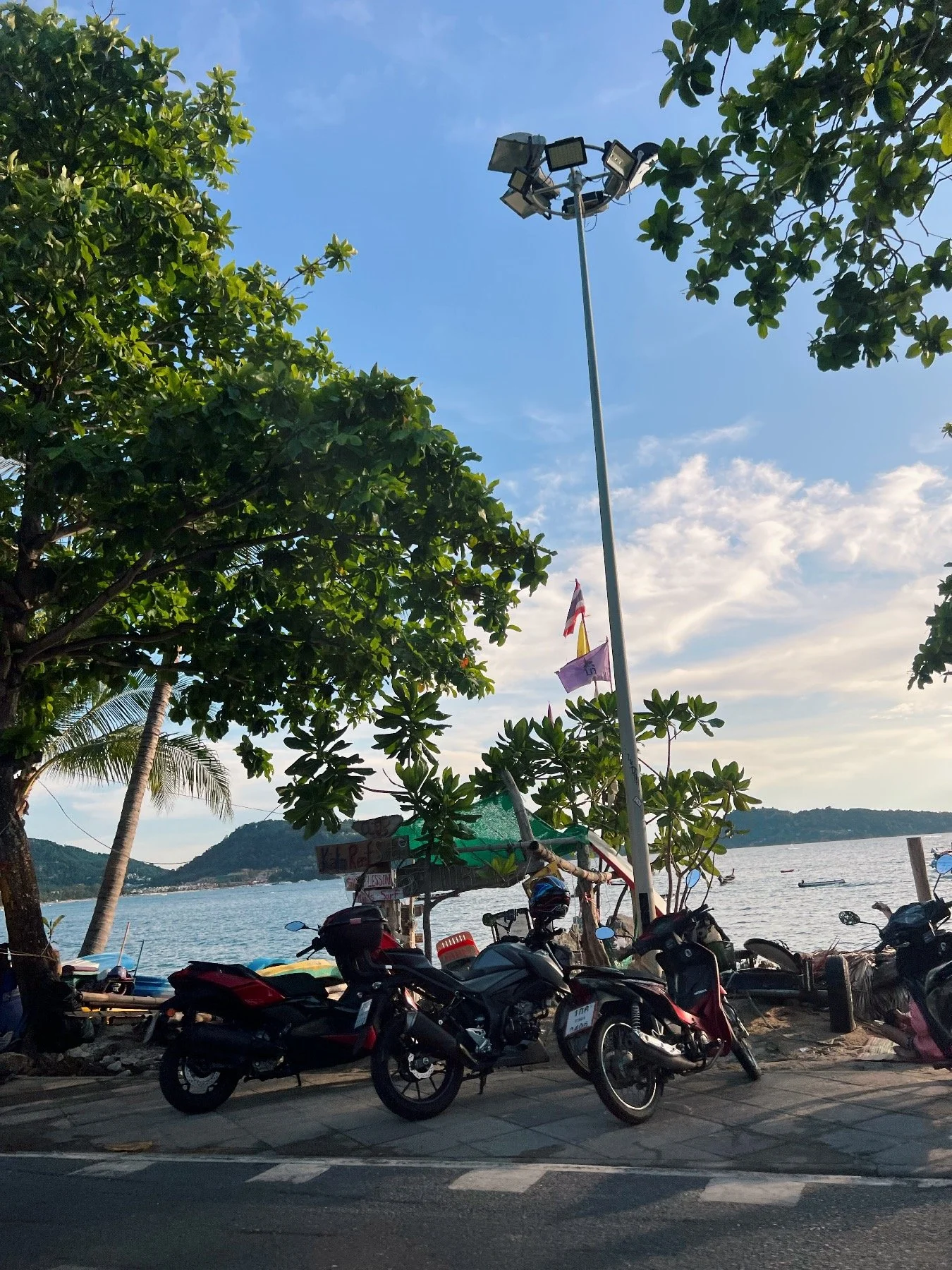 Scooters parked on the sidewalk near trees, with a view of the water and hills in the background, under a partly cloudy sky.