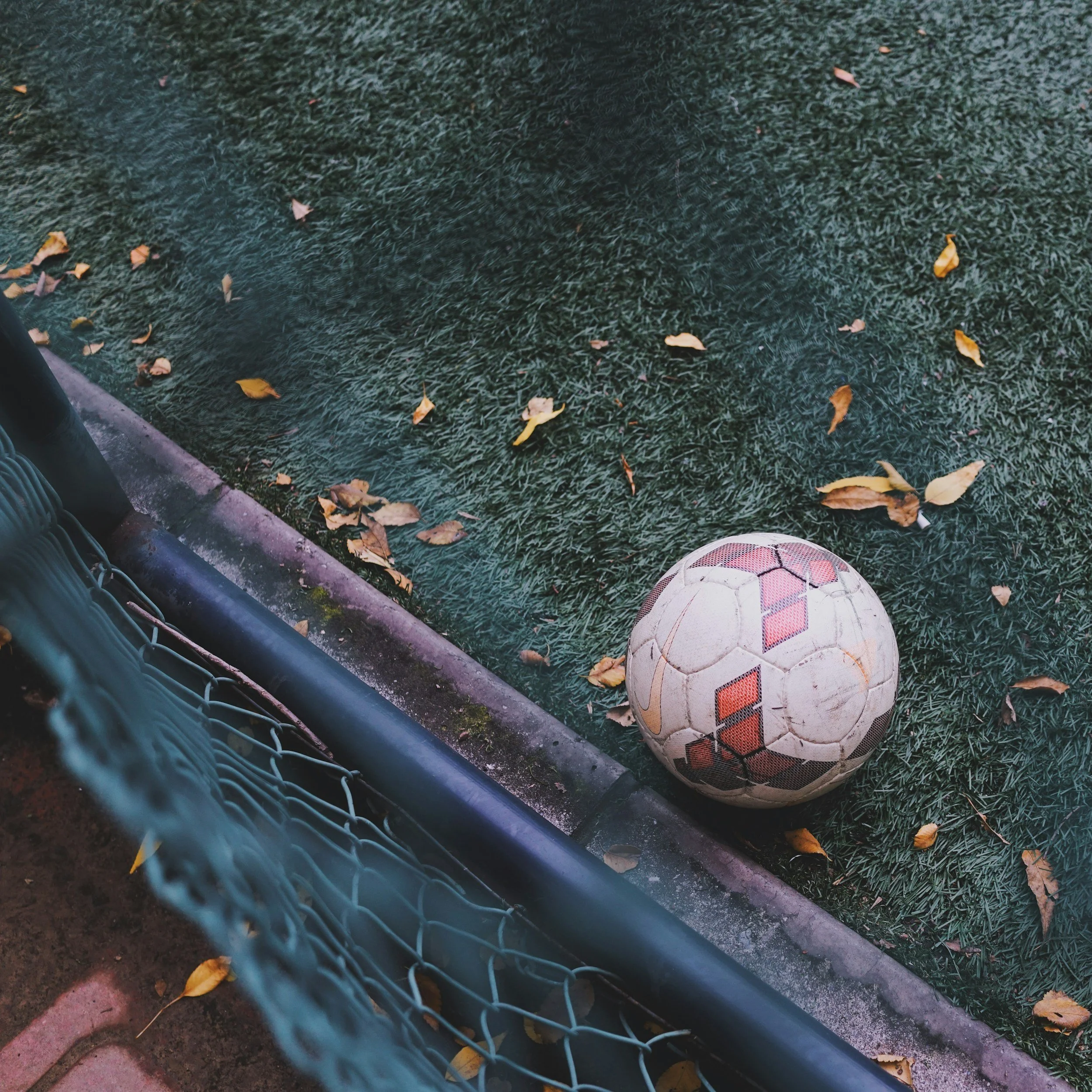 Soccer ball on artificial turf with fallen leaves and a chain-link fence nearby.