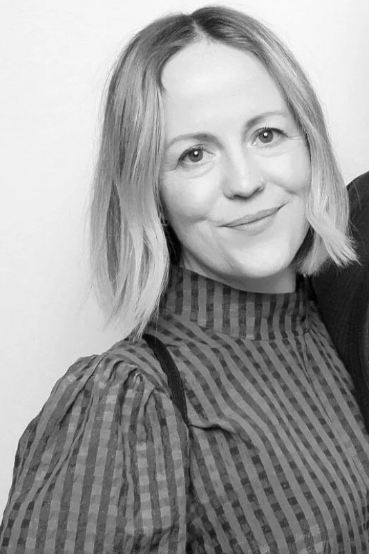 Black and white photo of a woman with shoulder-length light-colored hair, wearing a striped top, smiling at the camera.