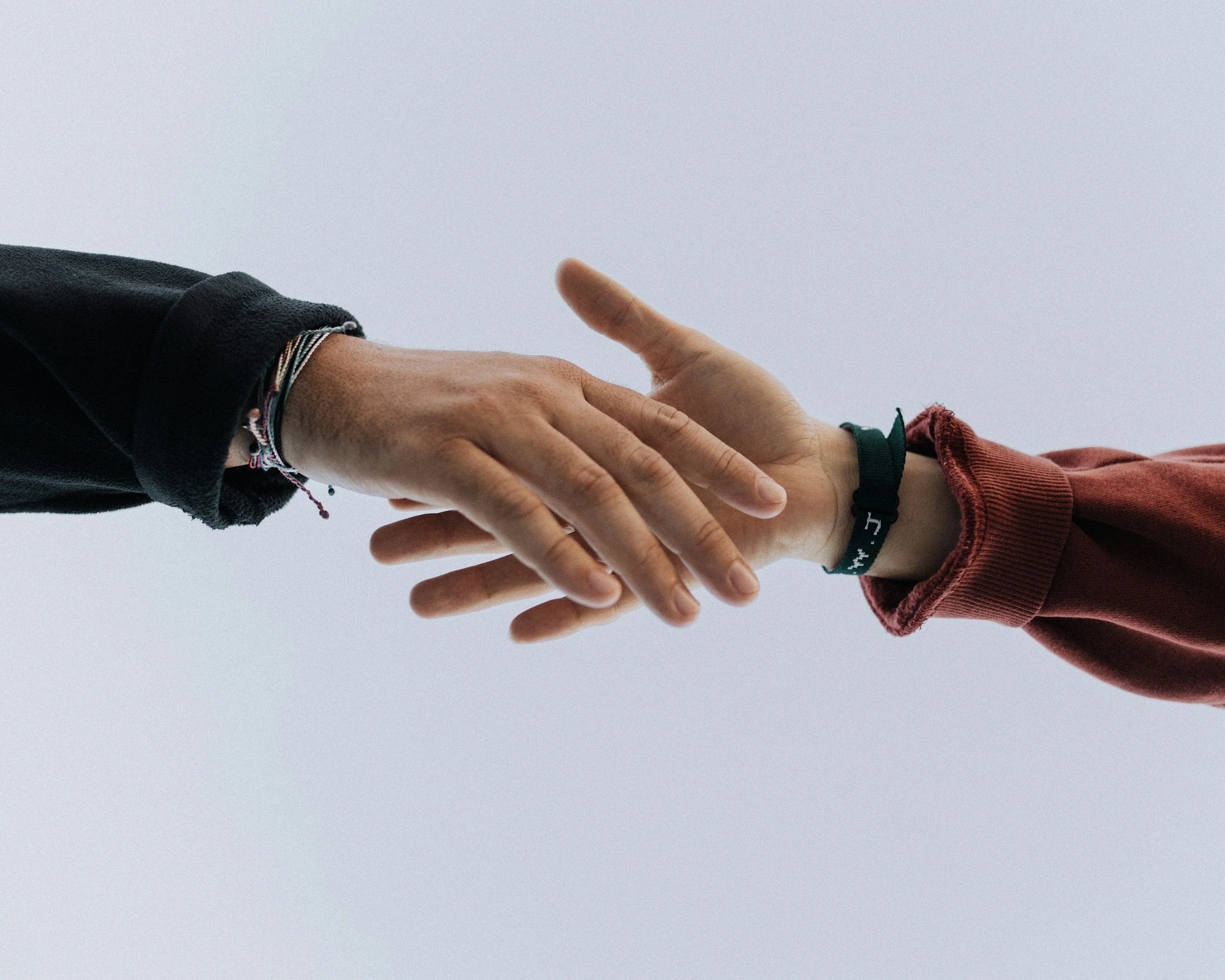 Two hands reaching towards each other, one with a black jacket and bracelets, the other with a red sleeve and a black wristband, against a plain gray background.
