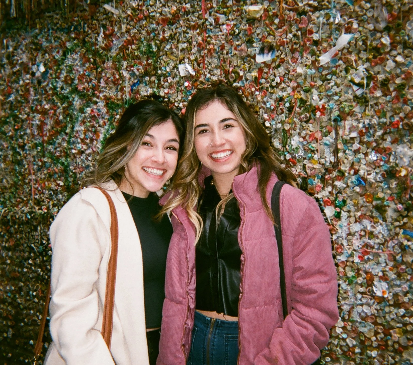 Two women smiling and posing for a photo in front of a large, textured wall covered with colorful debris, shells, and small objects.