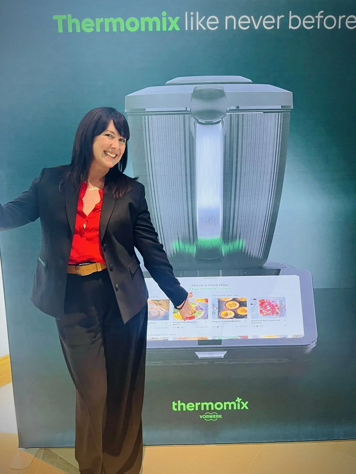 Woman in black blazer and red shirt pointing at a digital display with a large image of a Thermomix kitchen appliance and a touchscreen showing recipes, with the text 'Thermomix like never before' and 'thermomix' branding.