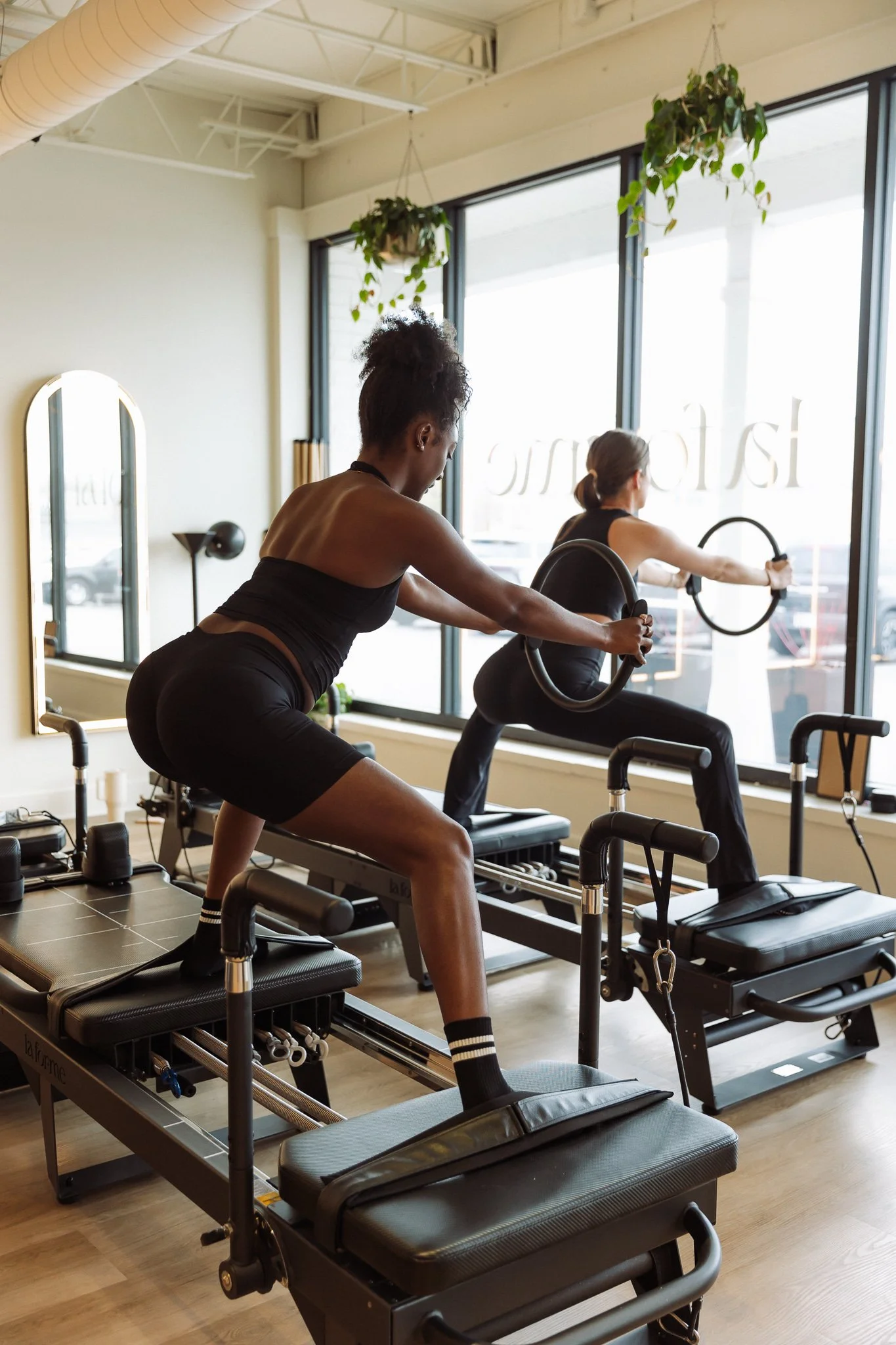 Two women working out on a Pilates reformer machine in a fitness studio with mirrors and modern lighting.