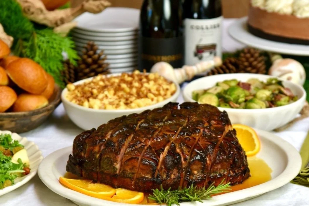A cooked prime rib roast on a white platter garnished with orange slices, surrounded by side dishes including salad, mashed potatoes, and vegetables, with wine bottles and a cake in the background.
