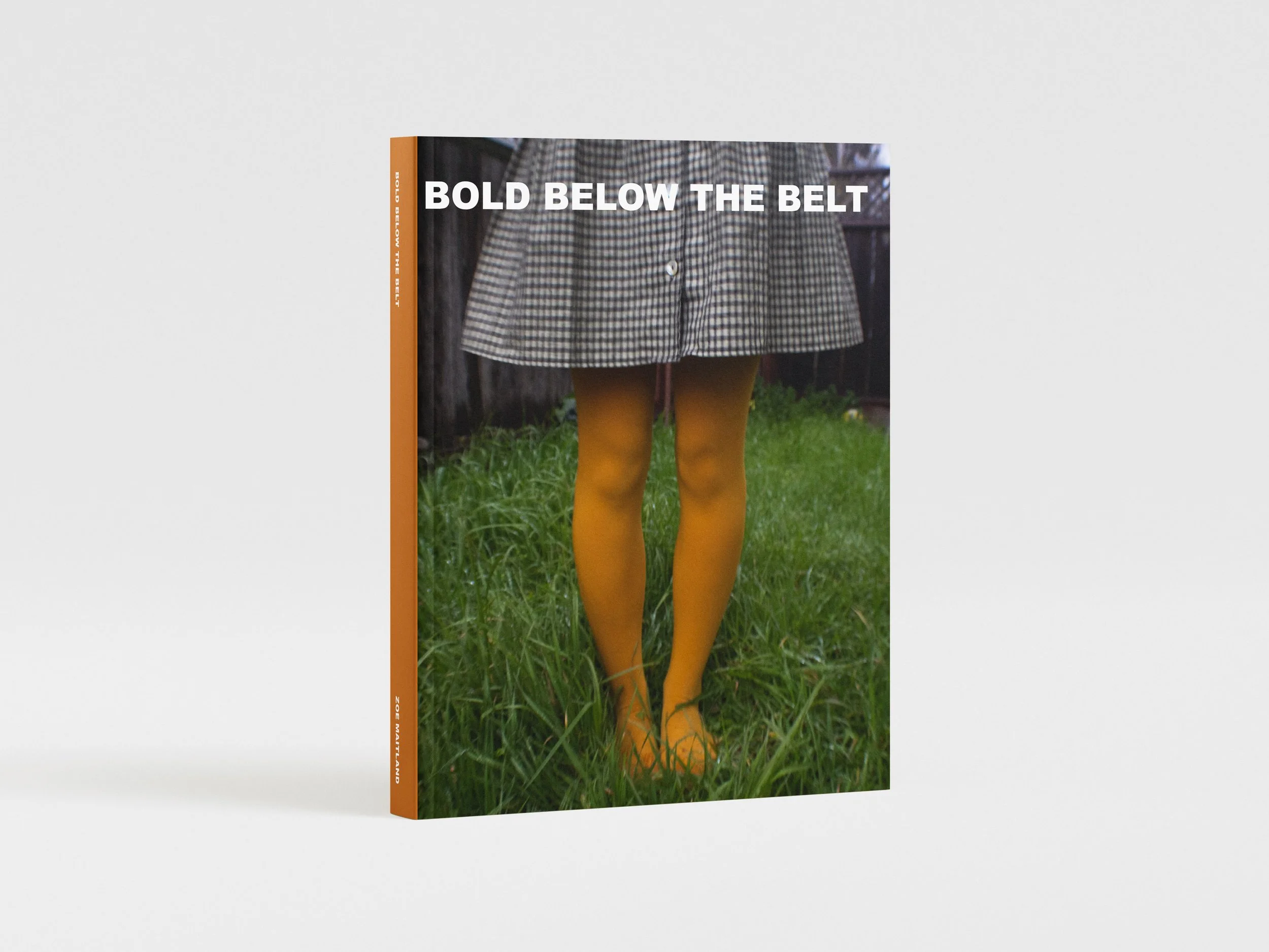 Book titled "Bold Below the Belt" showing a person in a checkered dress and yellow tights standing on grass.