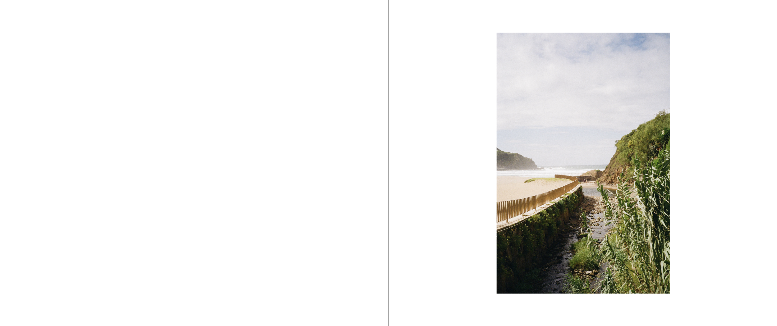 Scenic view of a sandy beach with a wooden railing, green vegetation, rocky cliffs, and ocean waves under a partly cloudy sky.
