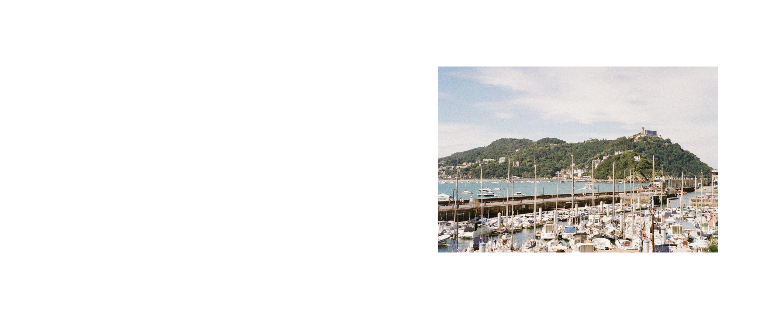 Marina with boats docked, harbor walls, hilly landscape in the background, and partly cloudy sky.