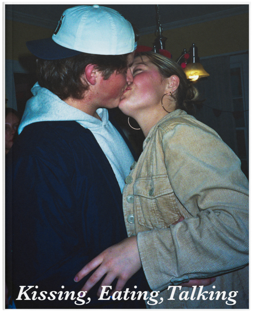 A young couple sharing a kiss indoors, with the girl wearing a beige jacket and large hoop earrings, and the boy wearing a gray hoodie and a baseball cap backwards, in a cozy setting with warm lighting.
