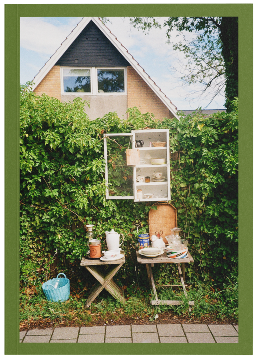 Outdoor scene of a garden with a green border, showing an old wooden table with dishes, a pitcher, and a cup, and a second table with various items, set against a background of a leafy hedge and a house with a triangular roof and large window.