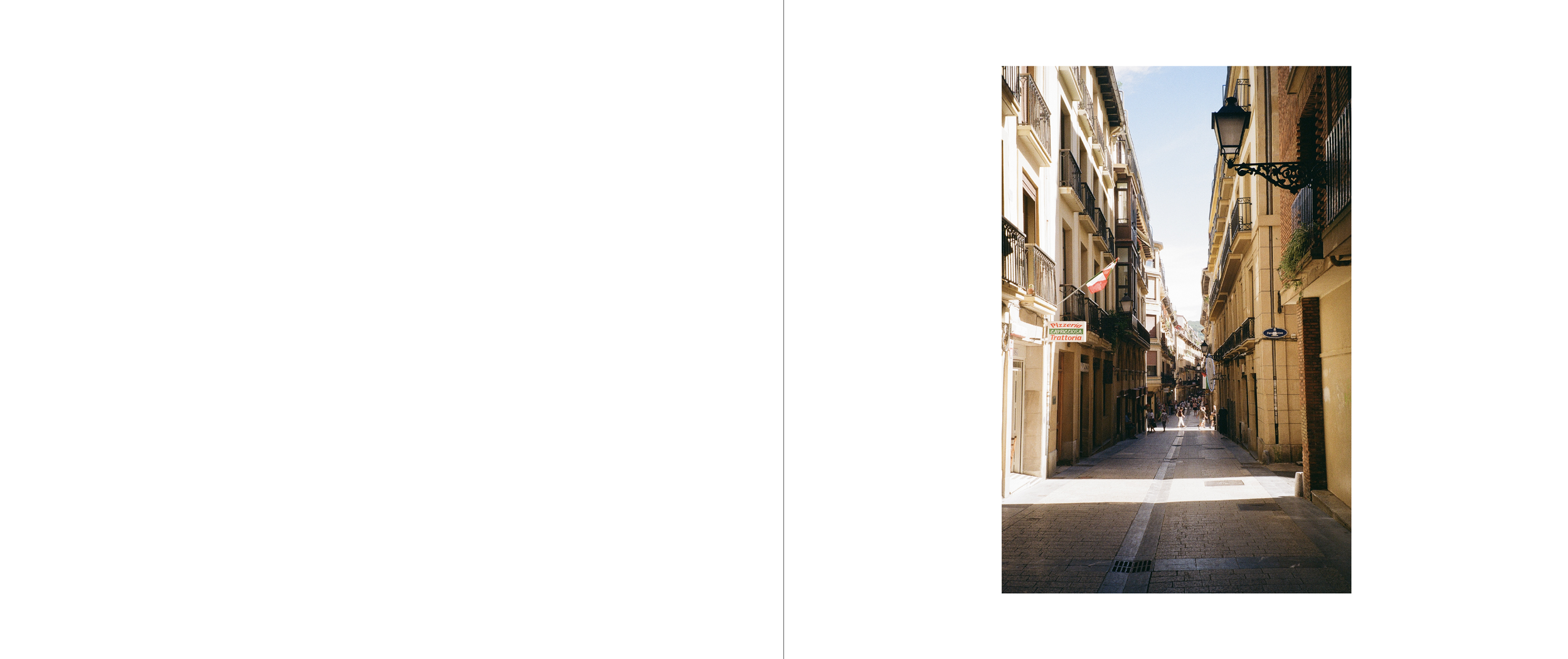 A narrow city street lined with multi-story buildings, featuring balconies, street lamps, and a flag, with pedestrians walking along the sidewalk during daytime.