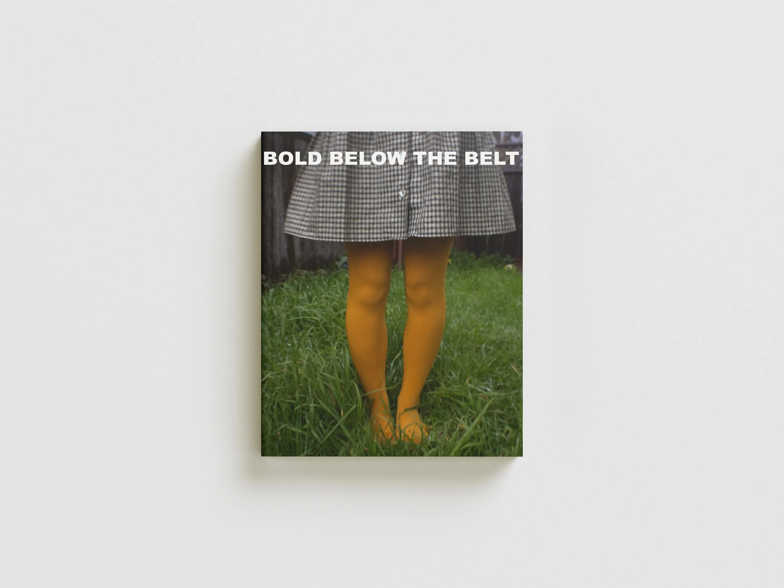 Book cover titled 'Bold Below the Belt' with a photo of a person's legs wearing yellow tights and a checkered skirt, standing on grass in a backyard.