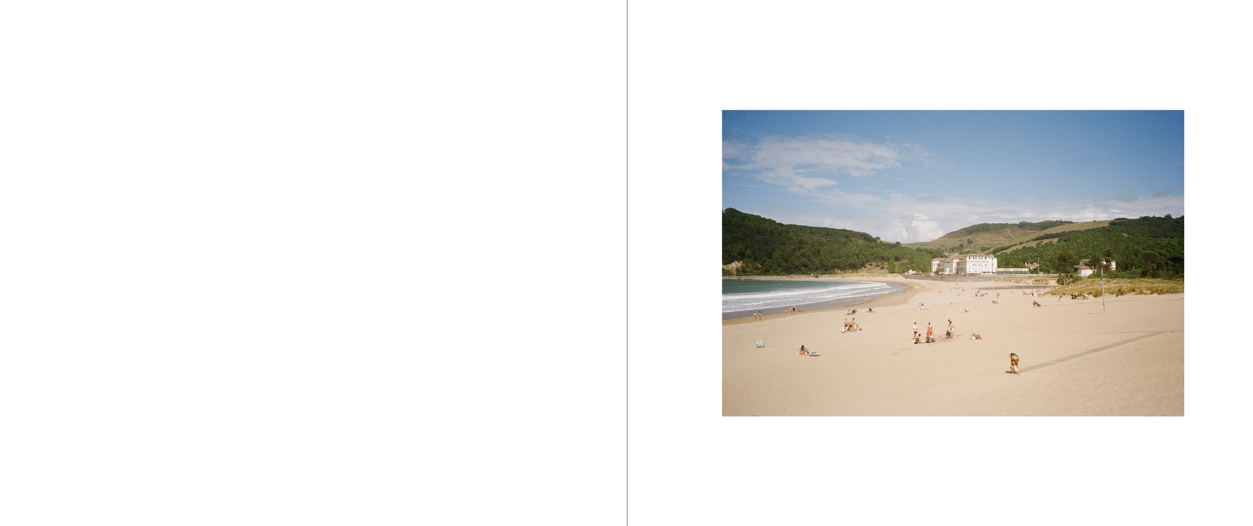 A sandy beach with people relaxing, walking, and sunbathing, backed by green hills and a blue sky with some clouds.