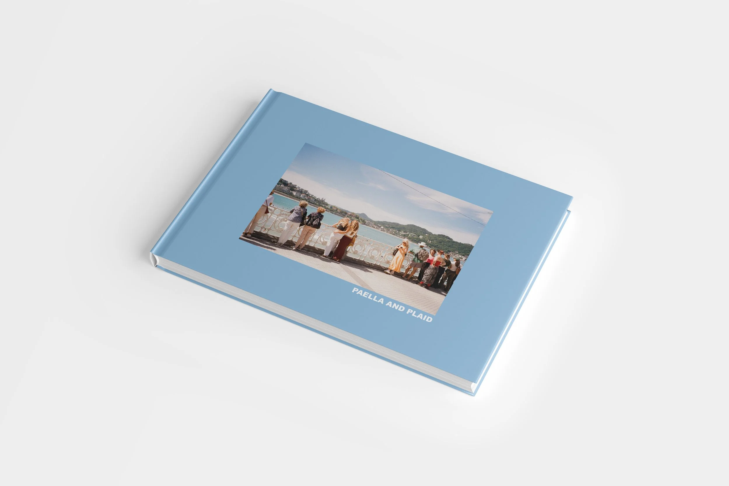 A light blue hardcover photo album titled 'Paella and Plaid' with a picture of people looking over a scenic waterfront view on the cover.