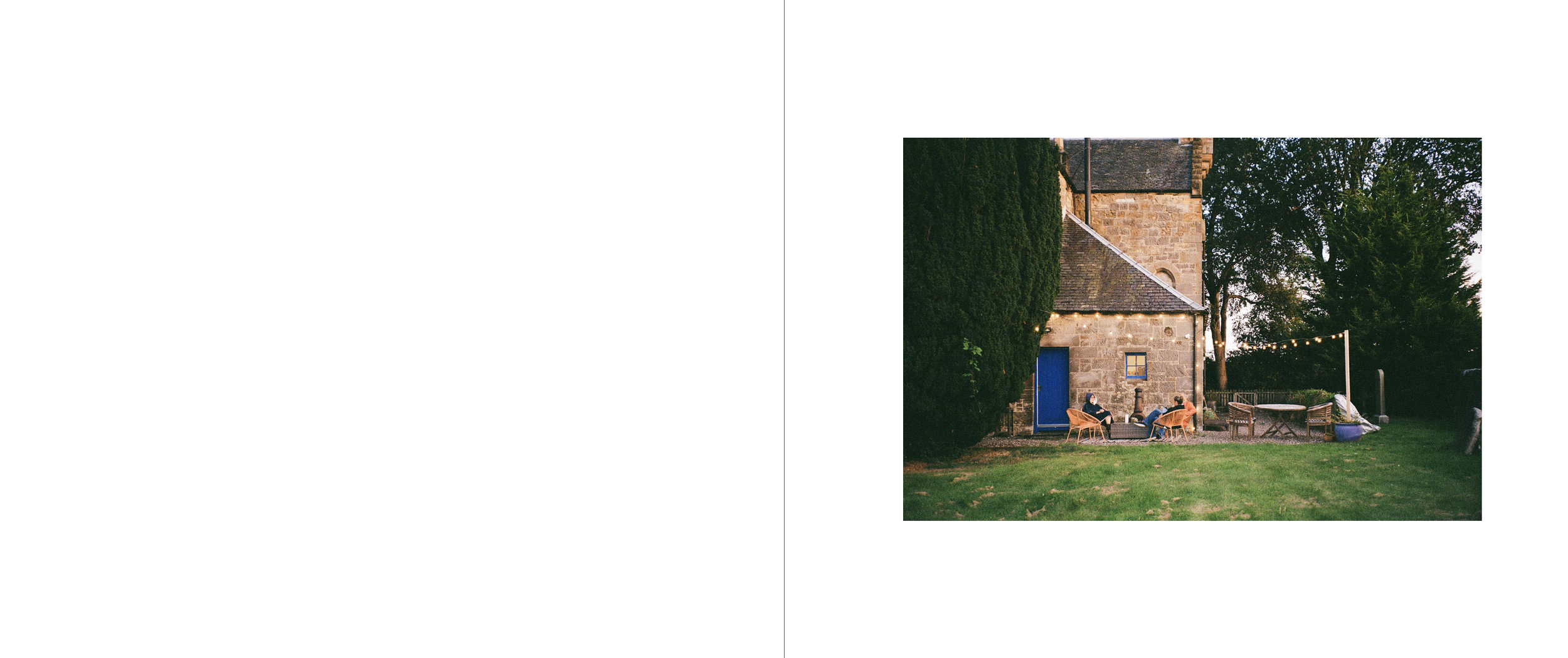 People sitting on outdoor chairs and sofa, illuminated string lights, in a backyard yard with trees and grass, adjacent to a stone building with a blue door.