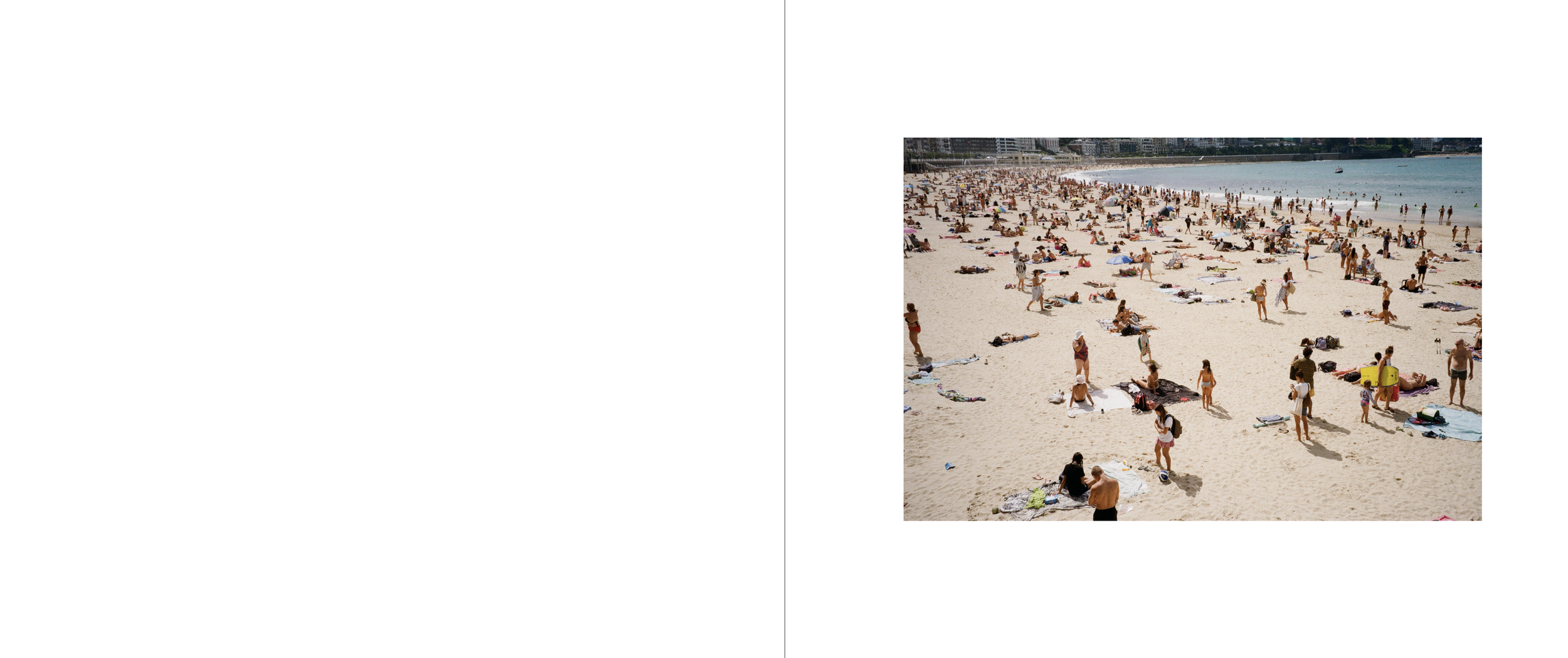 Crowded sandy beach with many people sunbathing, walking, and standing near the shoreline, with buildings in the background and the ocean to the right.