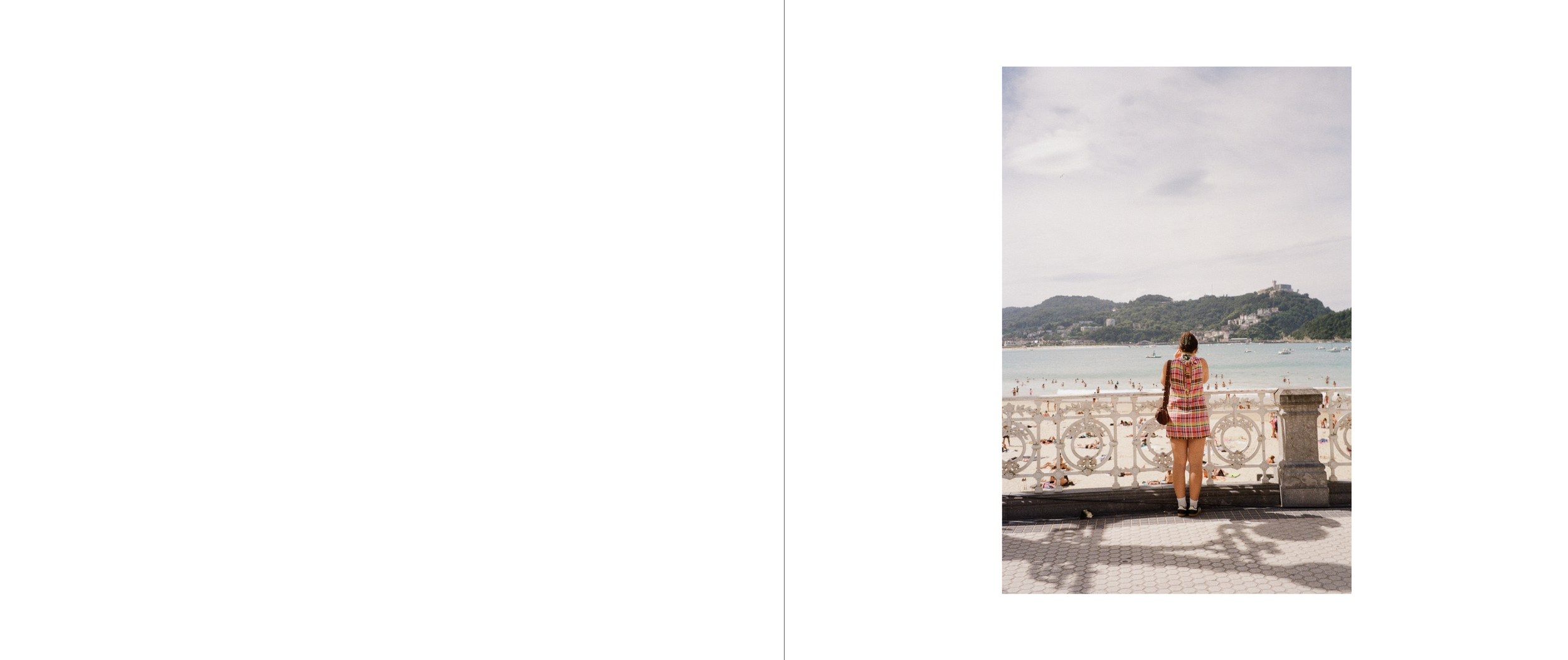 A woman stands by a rail overlooking a beach with people swimming and walking, with hills and buildings in the background.