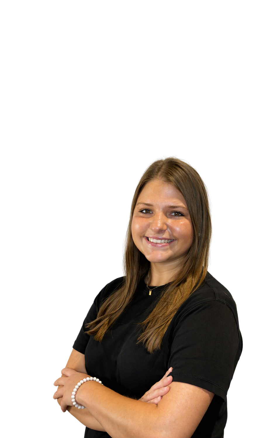 Young woman with long brown hair smiling, wearing a black t-shirt, white beaded bracelet, and a small pendant necklace, standing with arms crossed.