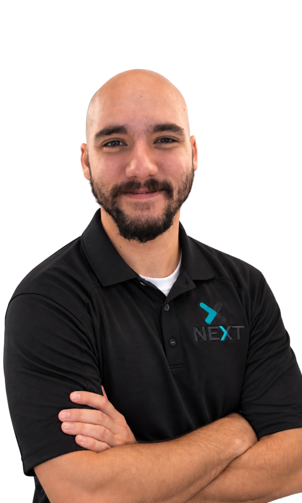 A smiling man with a beard and shaved head wearing a black polo shirt with the NEXT logo, arms crossed, standing against a black background.