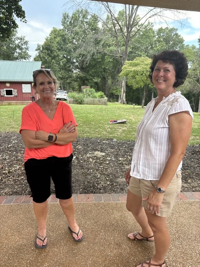 Two women standing outdoors on a patio, smiling and wearing casual summer clothes. One is in a red top and black shorts, the other in a white striped shirt and khaki shorts.