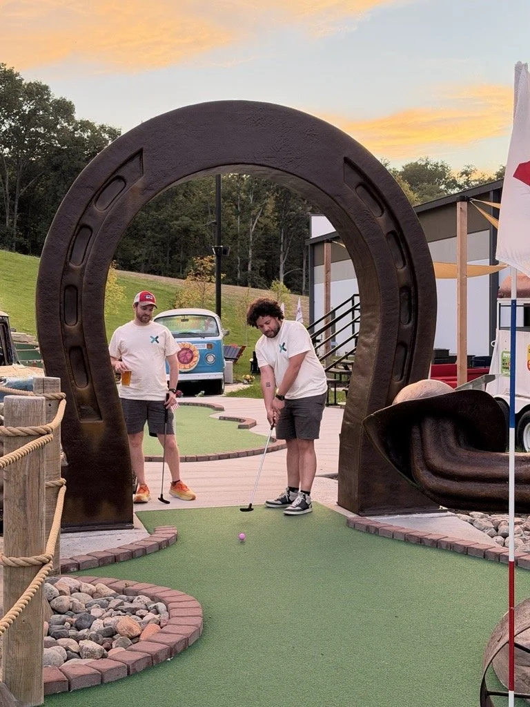 Two men playing miniature golf at an outdoor course during sunset. One is preparing to hit a pink golf ball with a putter while the other observes. The course has a tunnel obstacle with a large horseshoe shape and decorative rocks.