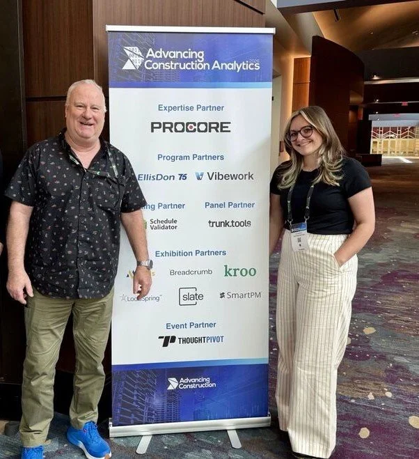 Man and woman standing next to an "Advancing Construction Analytics" conference banner with logos of various partner companies.