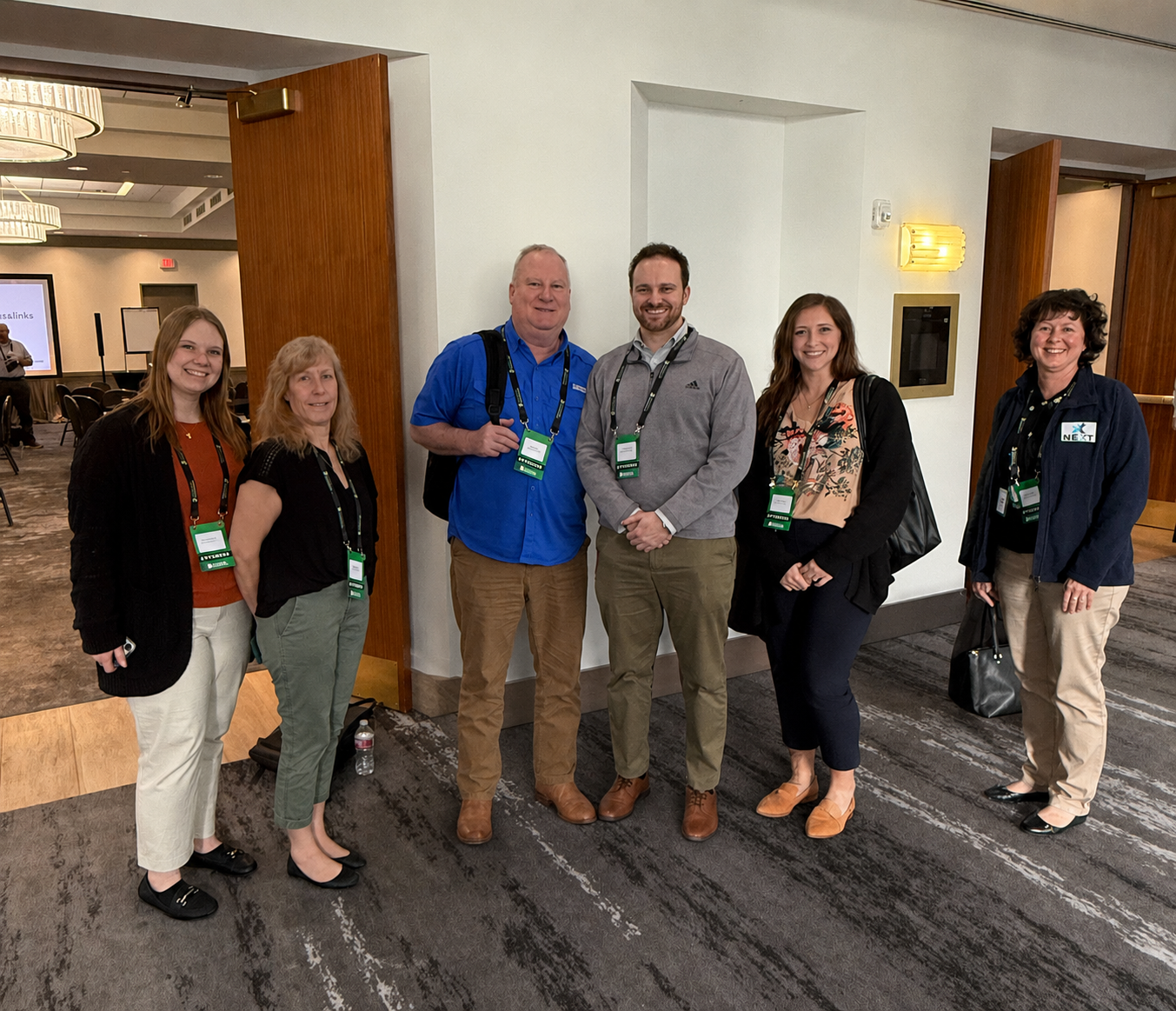 Group of six people standing indoors at a conference, smiling at the camera. They are wearing conference badges around their necks.