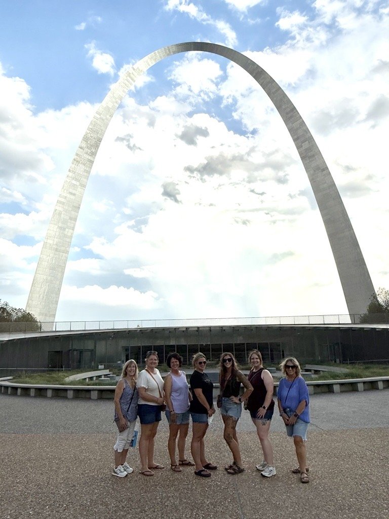 Group of seven women standing in front of the Gateway Arch in St. Louis, Missouri, with partly cloudy sky overhead.