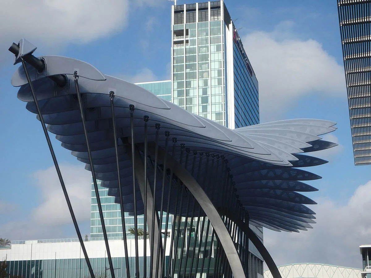 Modern sculpture resembling stylized wings with vertical supports, set in an urban area with tall buildings and a partly cloudy sky.
