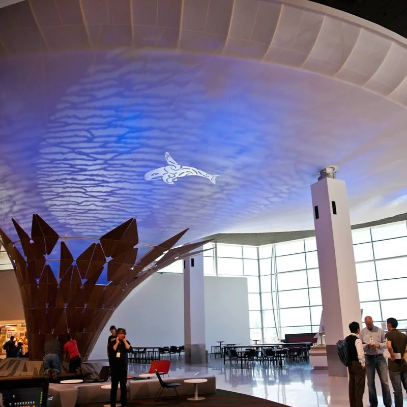 Interior of a modern building with a curved ceiling featuring blue lighting and a fish-shaped projection, large glass windows, and a few people talking near pillars and seating areas.