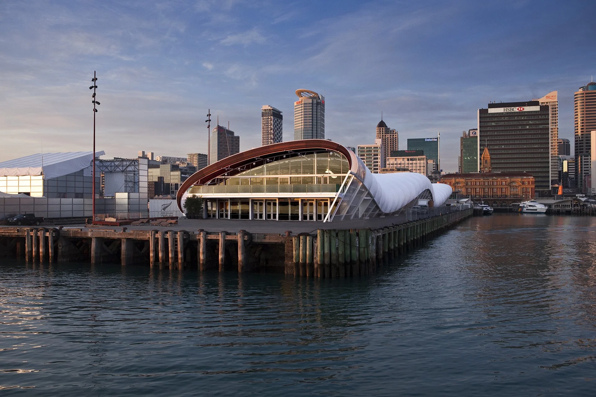 The Cloud, a modern waterfront building with a curved, white roof and glass facade, set against a city skyline with tall skyscrapers, under a partly cloudy sky at sunset.