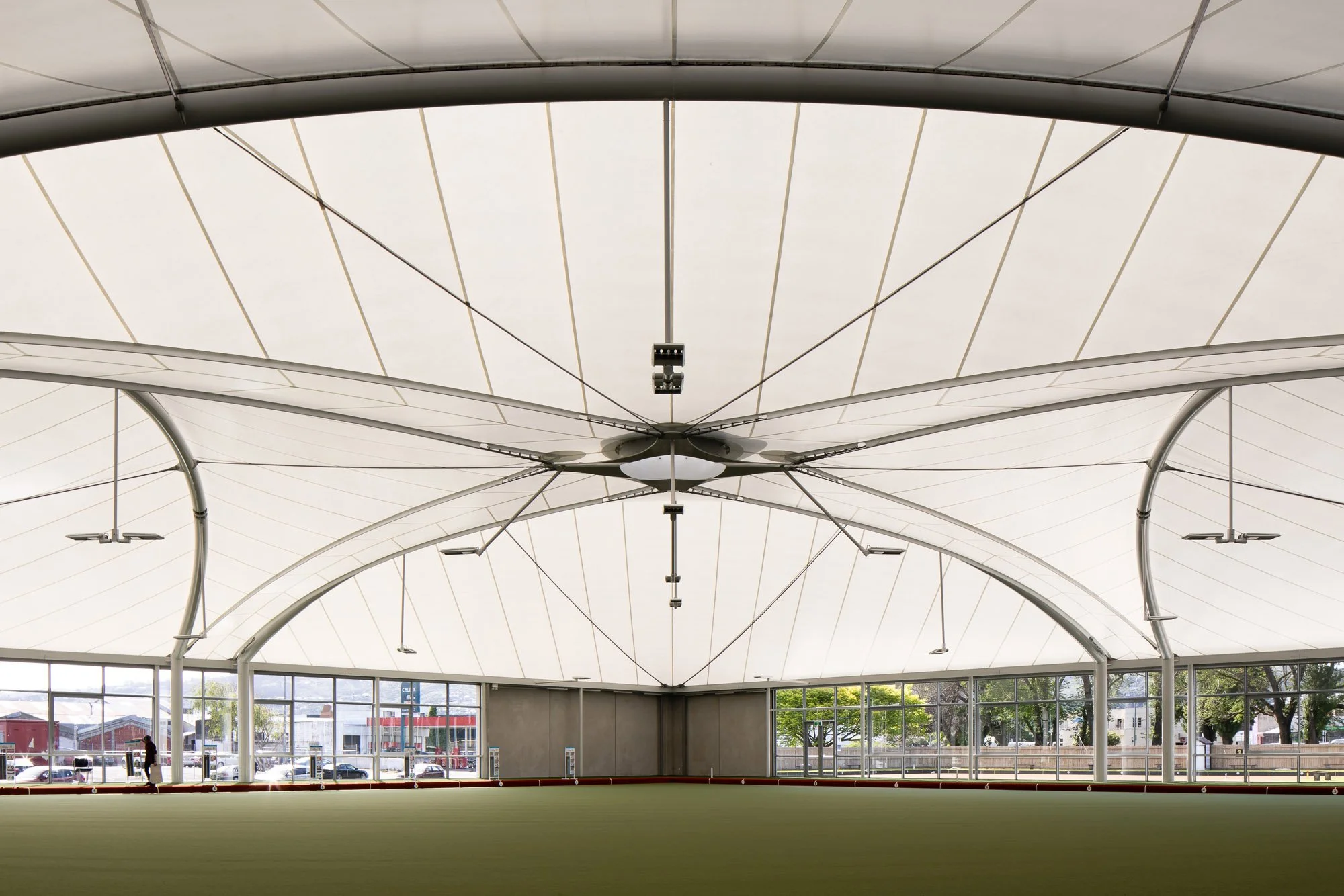 Interior view of a modern indoor sports facility with a curved fabric ceiling, large glass windows, and a grassy playing field.