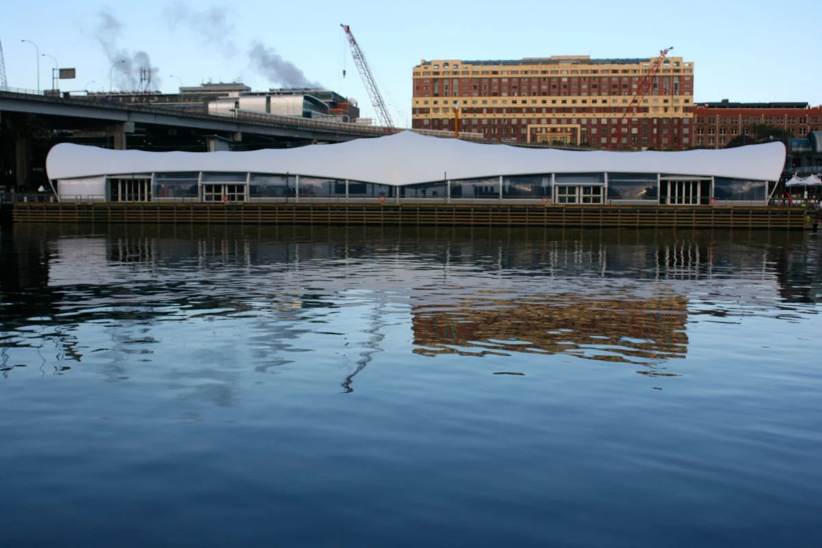A modern floating building with a white, wave-shaped roof on a body of water, with urban buildings and construction cranes in the background.