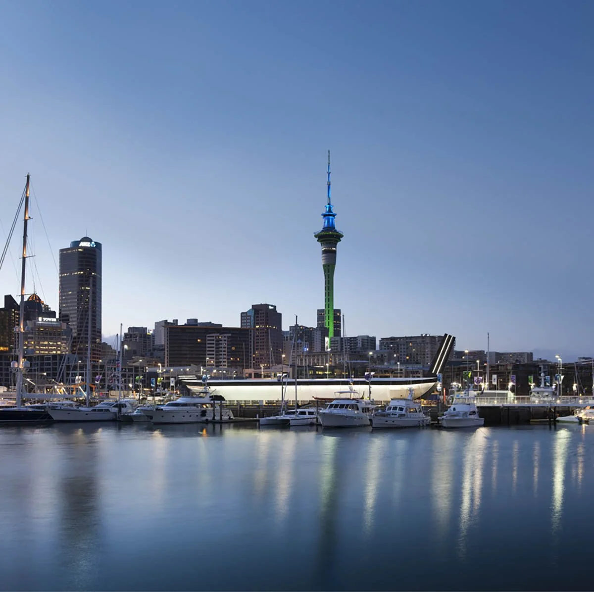 Night scene of Auckland waterfront with boats docked in the harbor and the Sky Tower illuminated in green and blue in the city skyline.