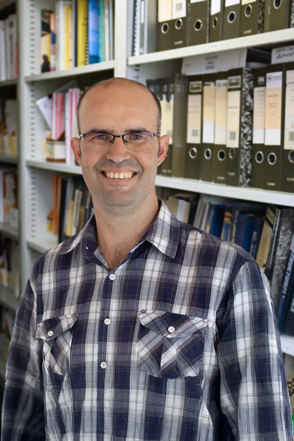 Hamish Scott-Davidson, Design Engineer. A smiling man with glasses and a plaid shirt standing in front of shelves filled with binders and books in an office setting.