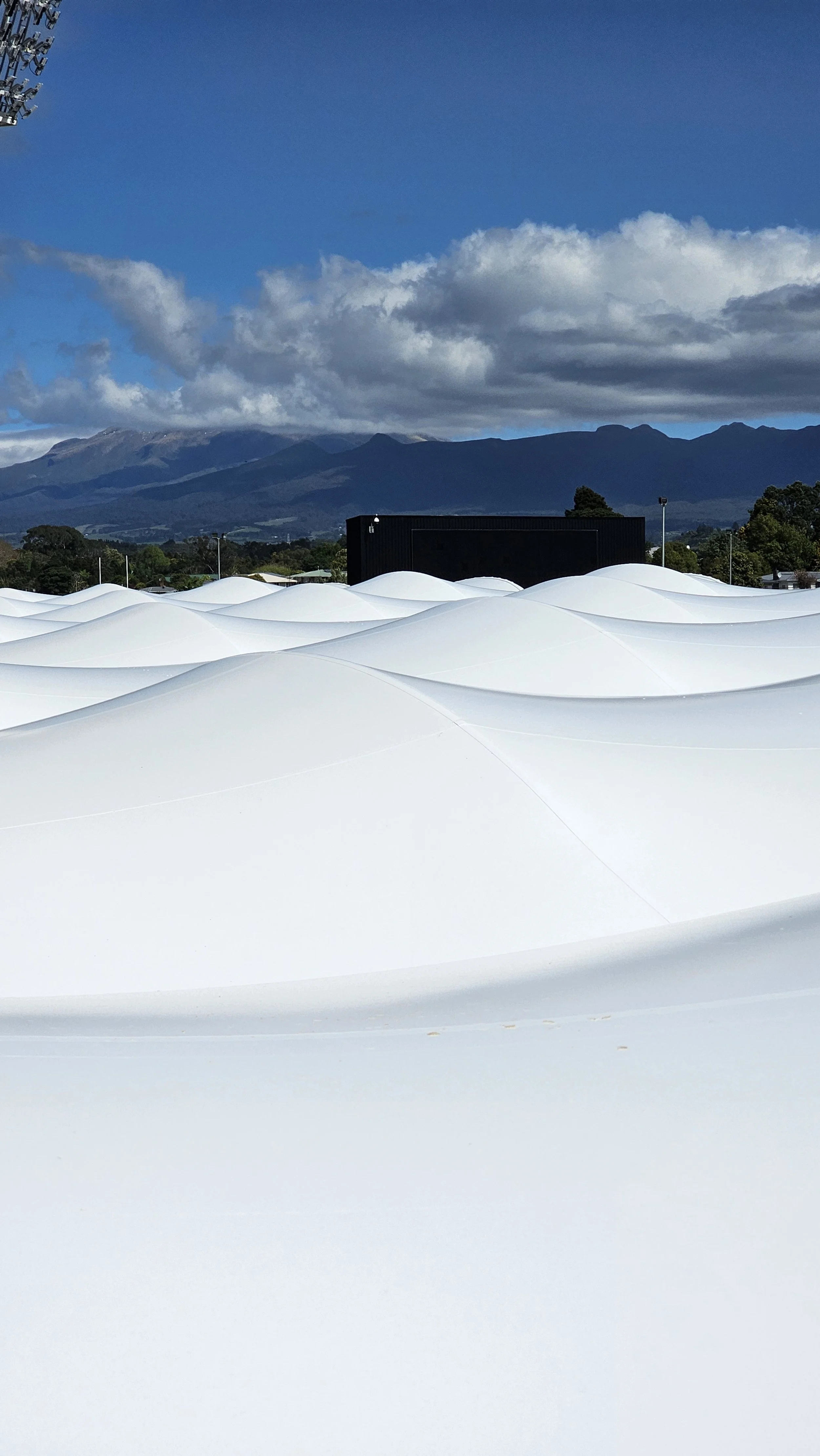 Across the rooves of Yarrow Stadium, Taranaki. White modern architectural structure with smooth, flowing curves against a backdrop of green trees, mountains, and a partly cloudy blue sky.